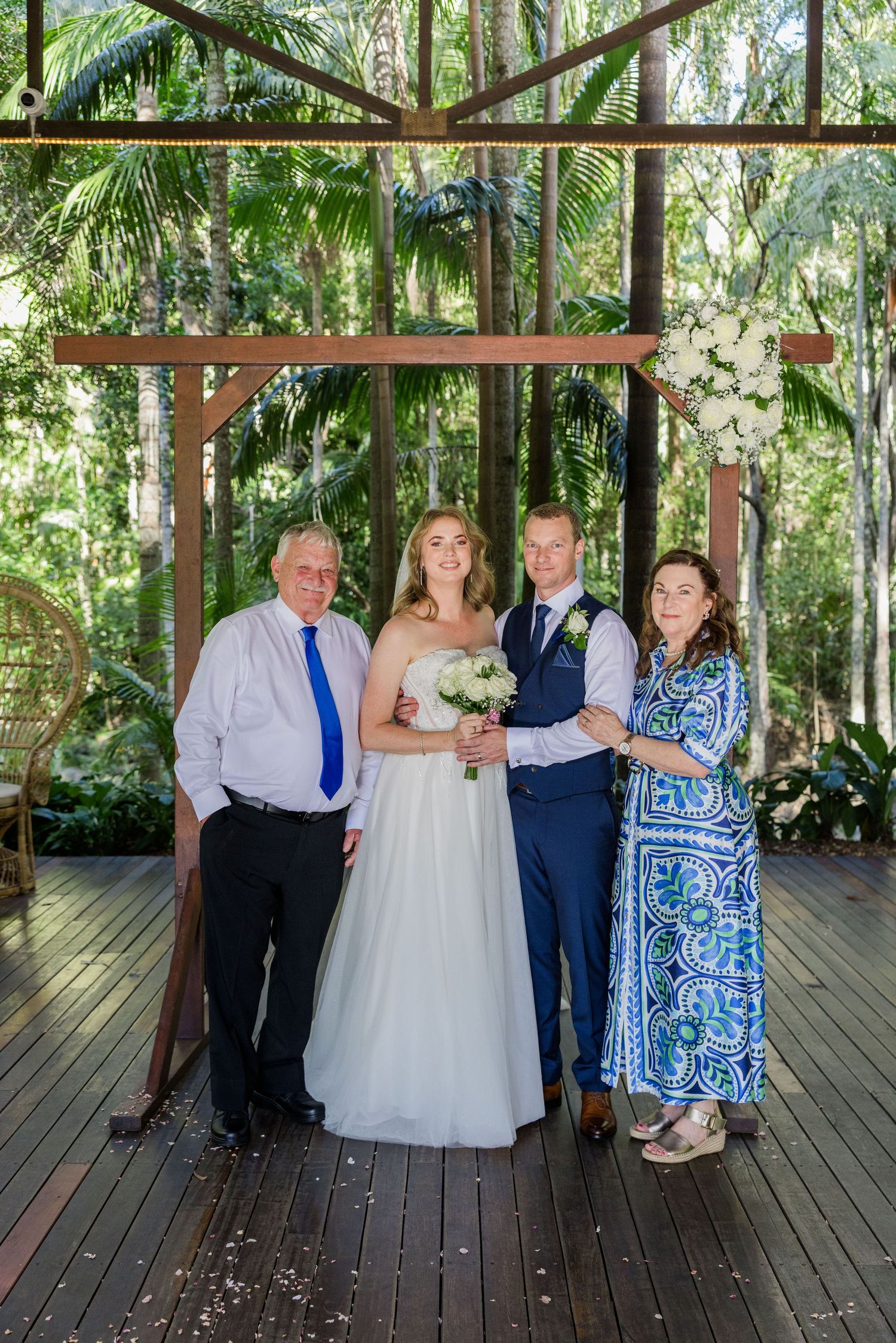 Wedding party poses together under a wooden pergola surrounded by lush tropical greenery and palm trees.