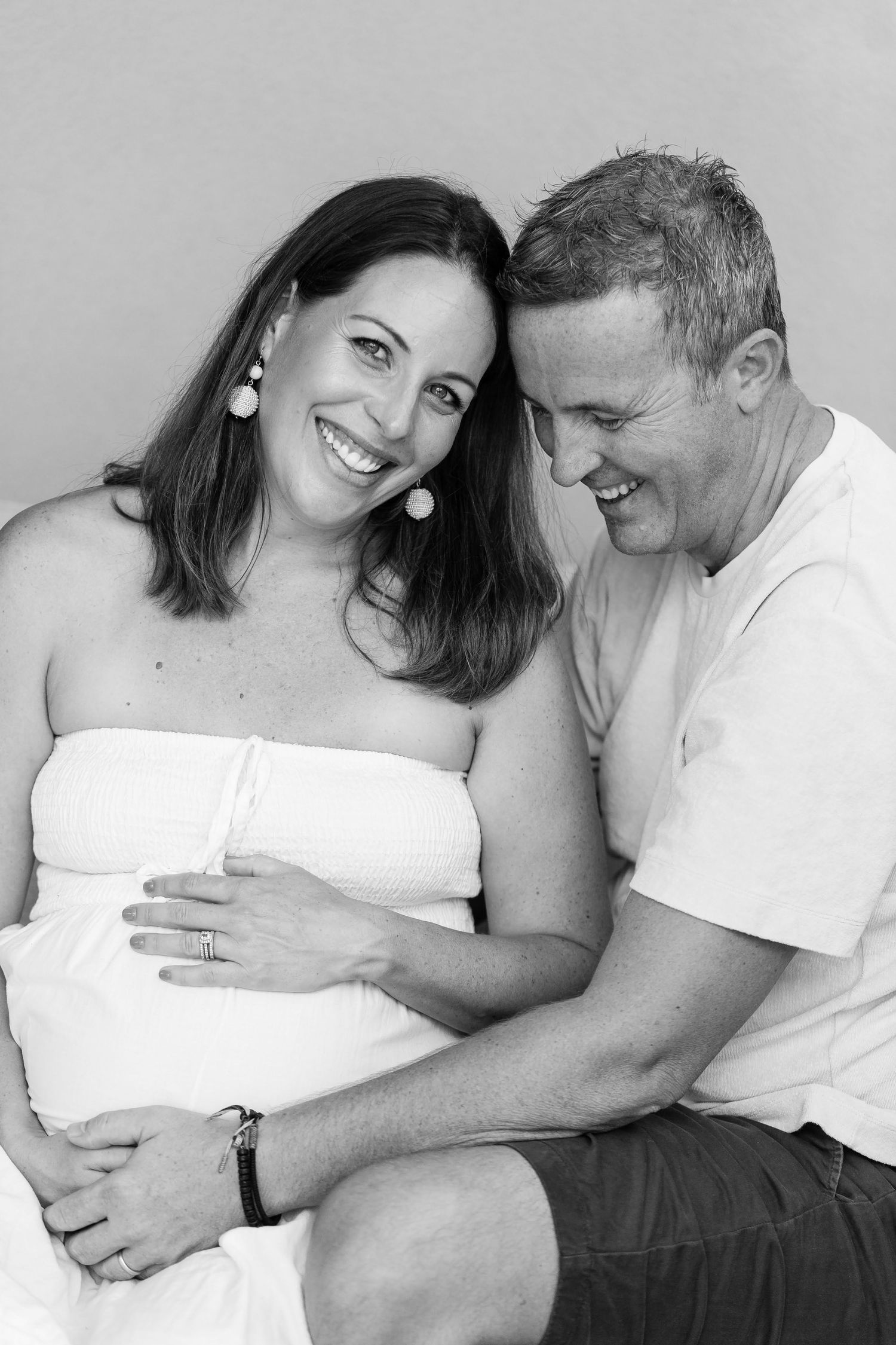 A black and white portrait shows an intimate moment between an expecting couple in casual attire.