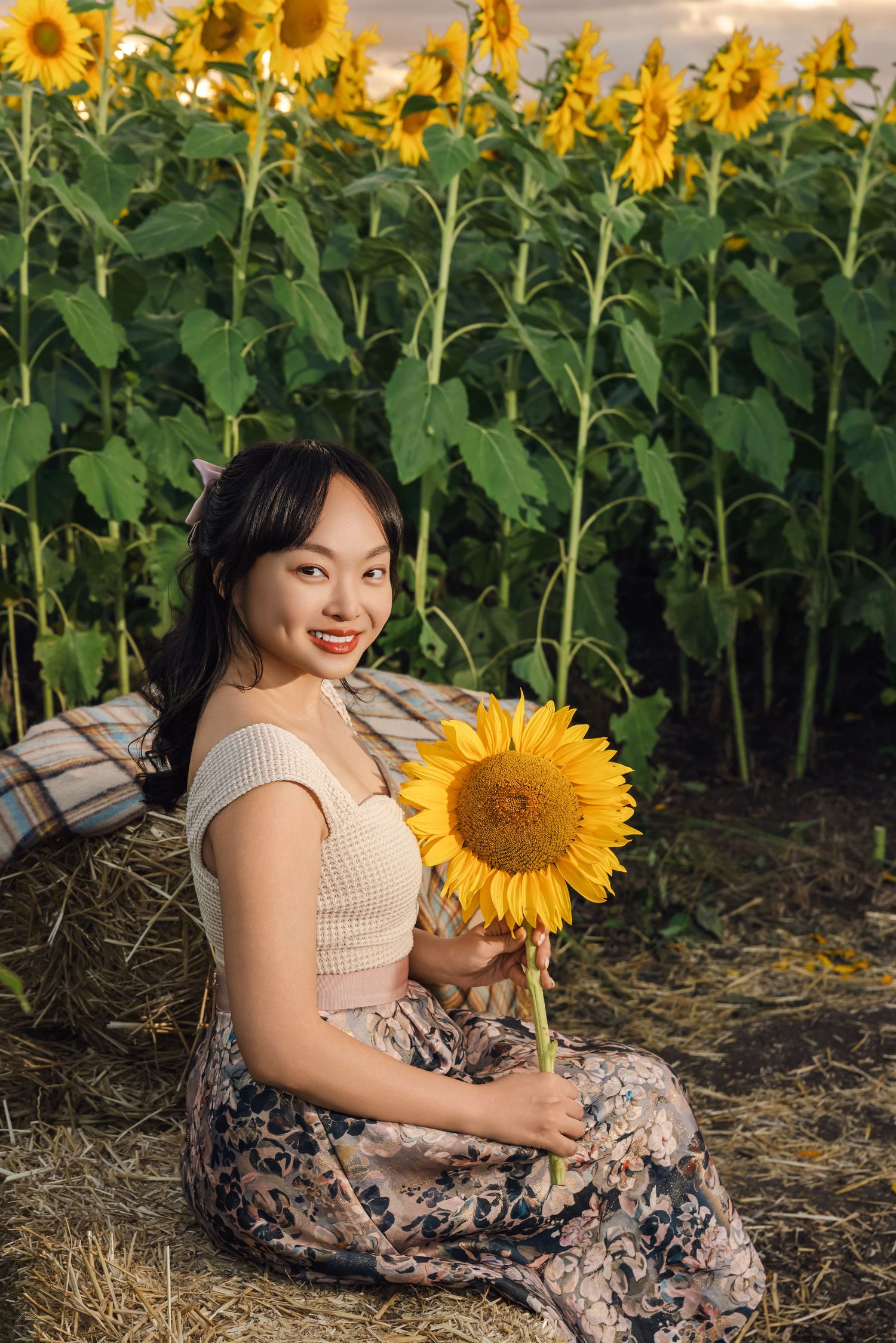 Smiling young woman sitting on hay bales holding a sunflower in front of a lush sunflower field.