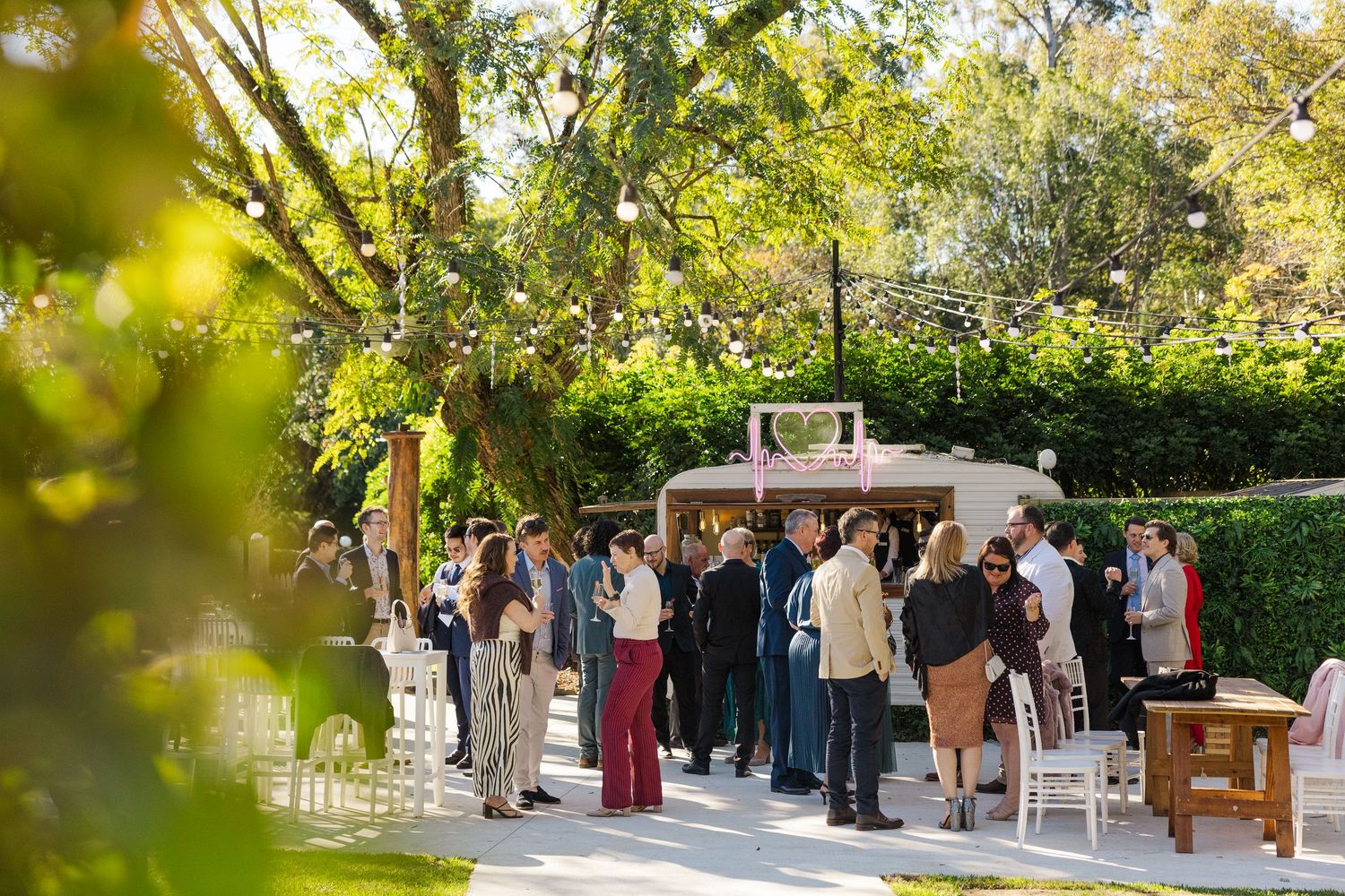 Guests mingle outdoors at an elegant garden wedding reception under lush green trees on a sunny day.