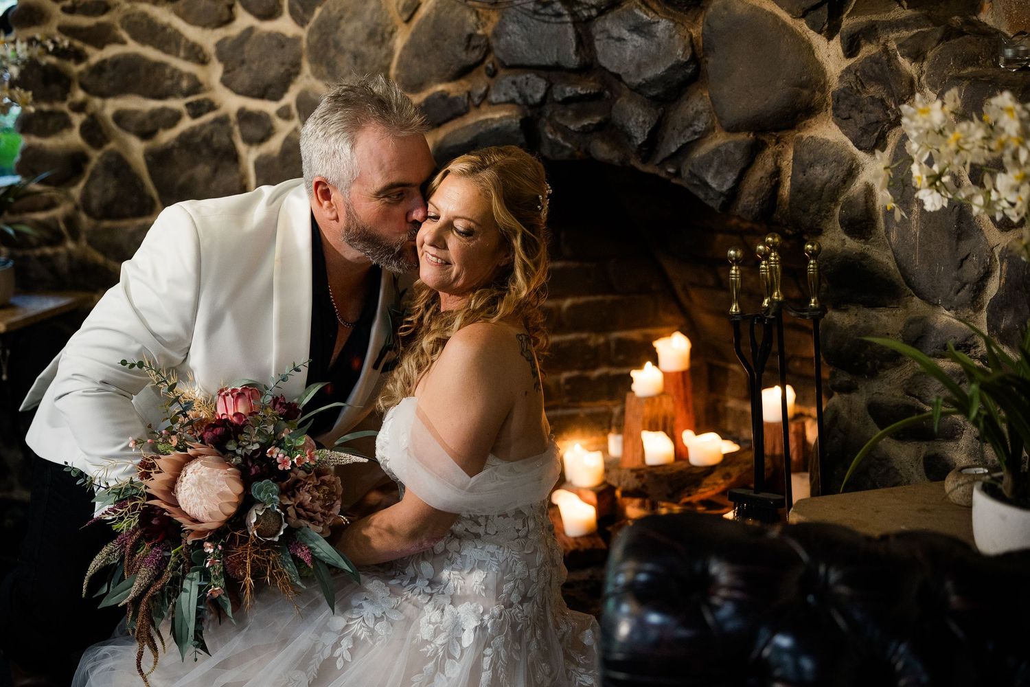 Bride and groom share a romantic kiss by a candlelit stone fireplace, bride holding a boho floral bouquet.