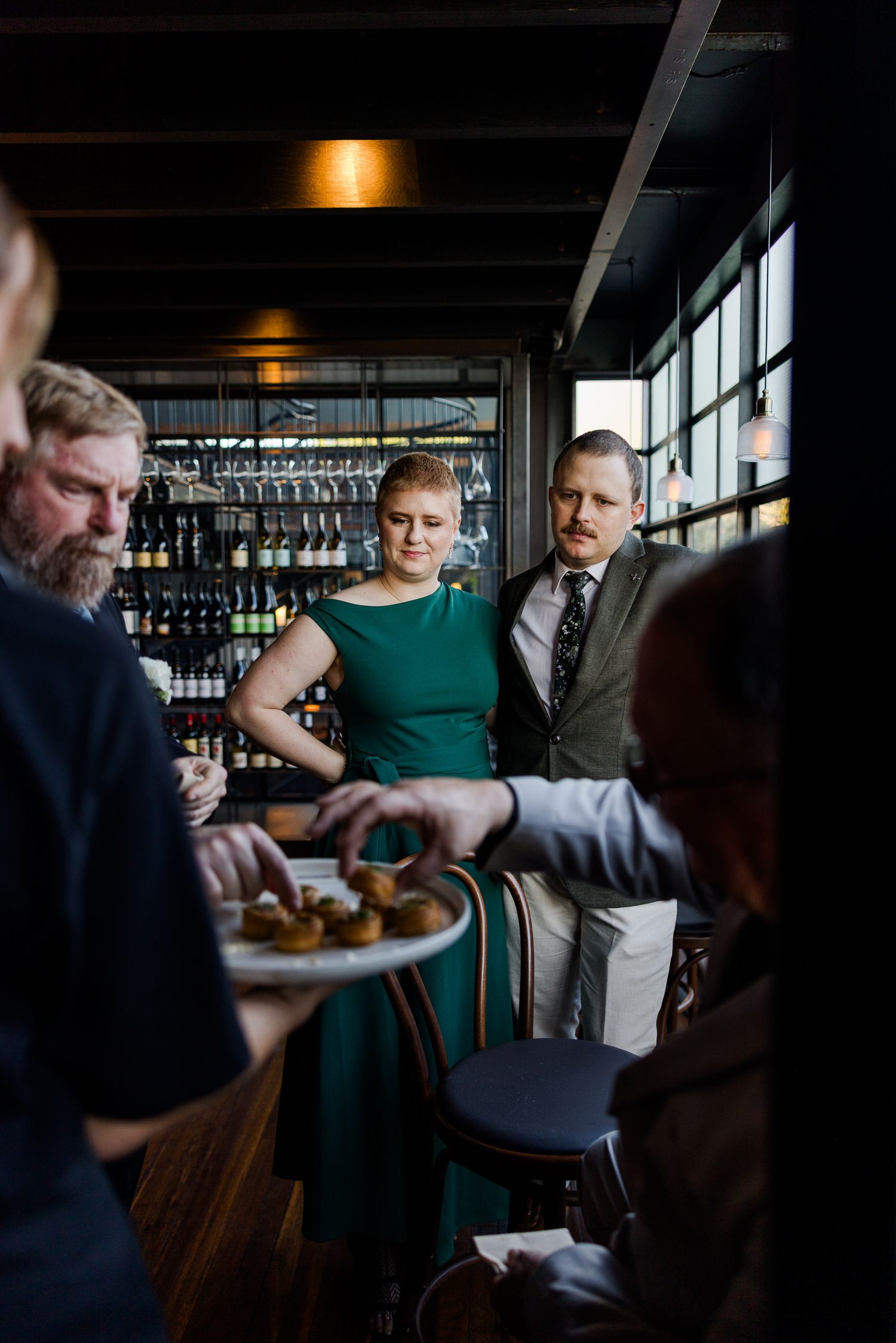 Guests at an upscale bar reception, server offering appetizers to elegantly dressed attendees near a wine rack.