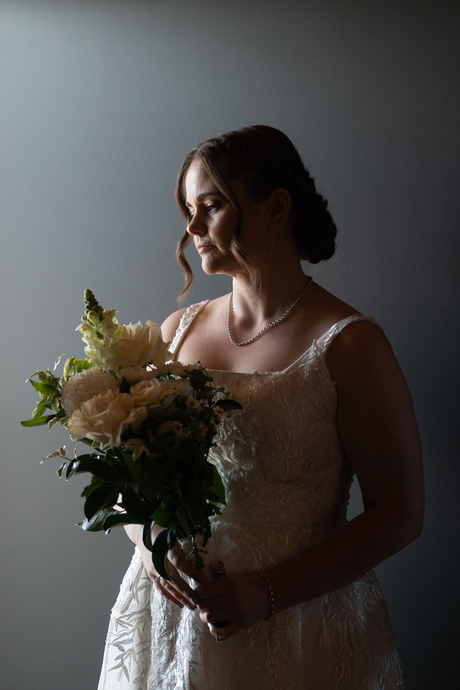 Bride in white lace gown holds cream floral bouquet, gazing softly to the side in a dramatic studio portrait.