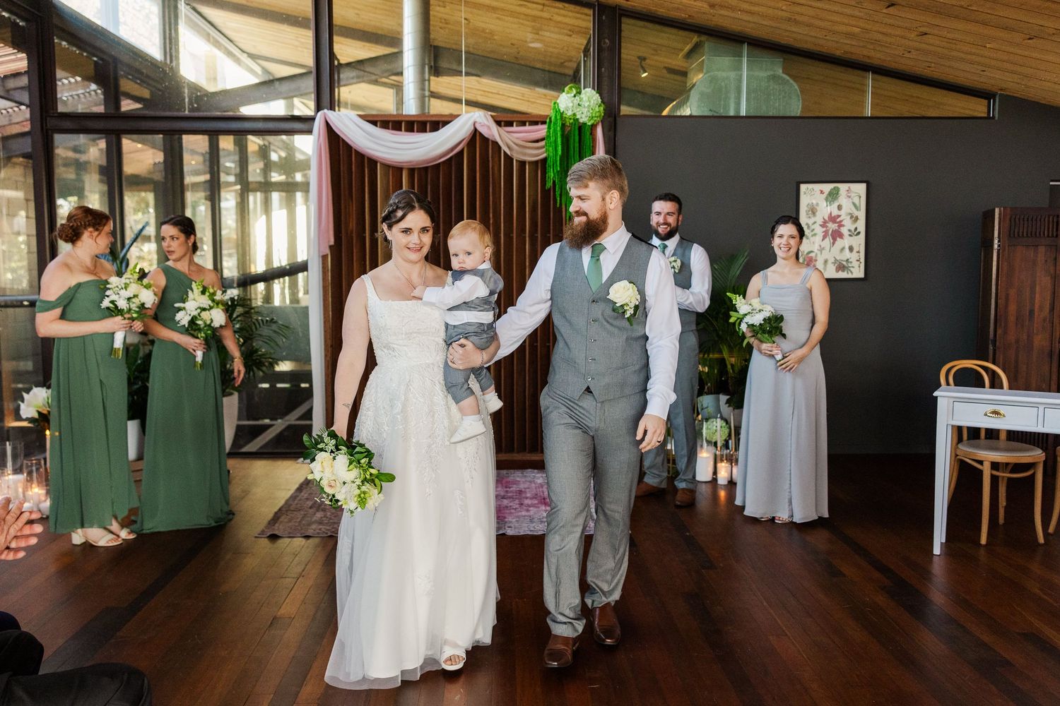 Bride and groom walk with baby after indoor wedding ceremony, bridesmaids in green and grey dresses stand nearby.