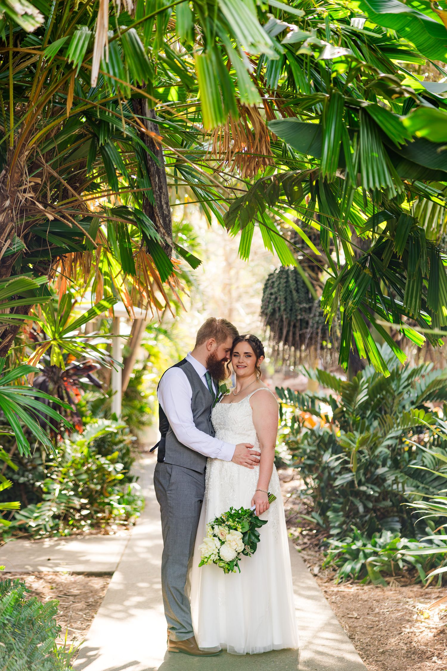 Bride and groom embrace on a garden path surrounded by lush tropical palms, bride holding white floral bouquet.