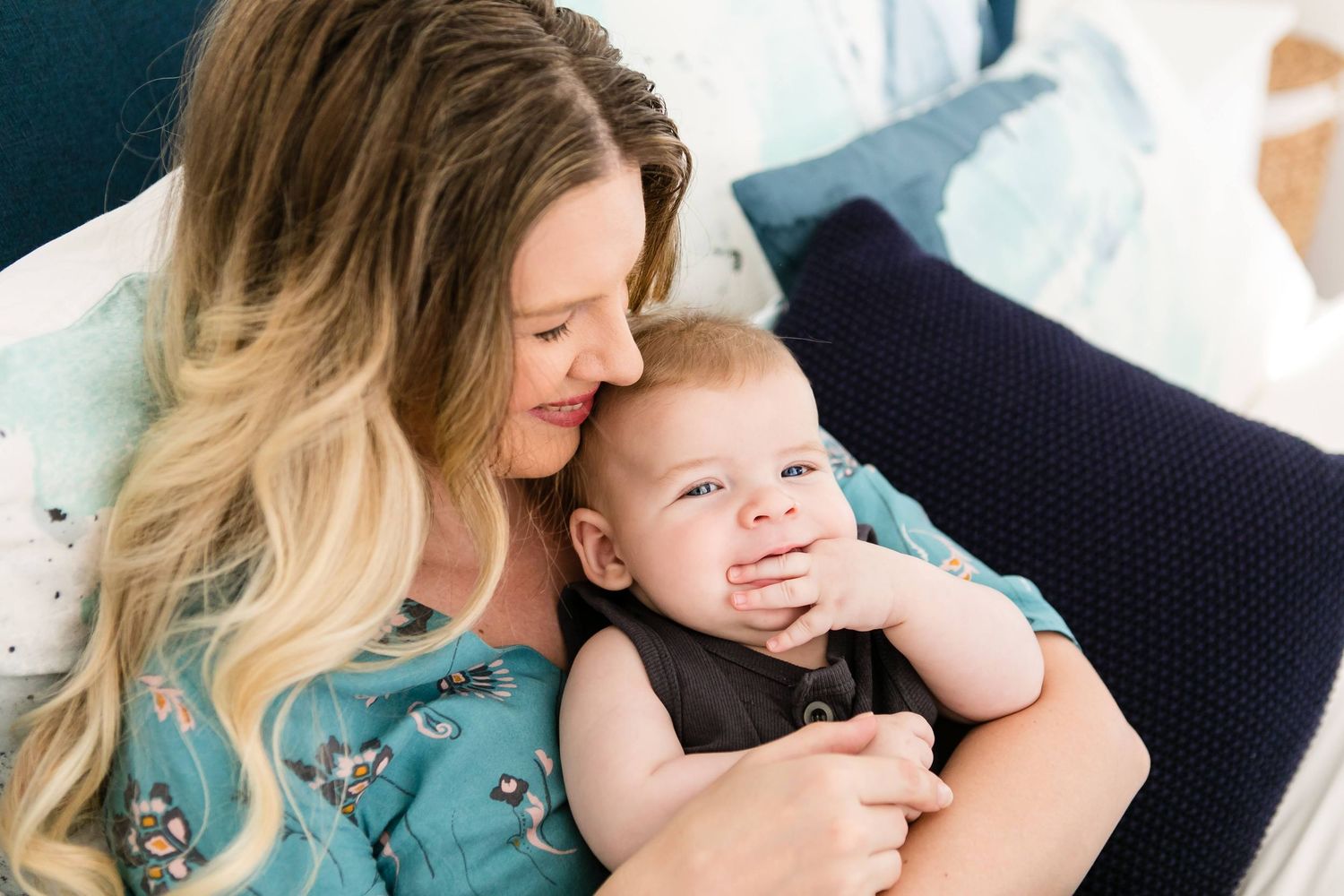 A loving mother with blonde hair smiles tenderly at her happy baby boy while relaxing on a cozy bed.