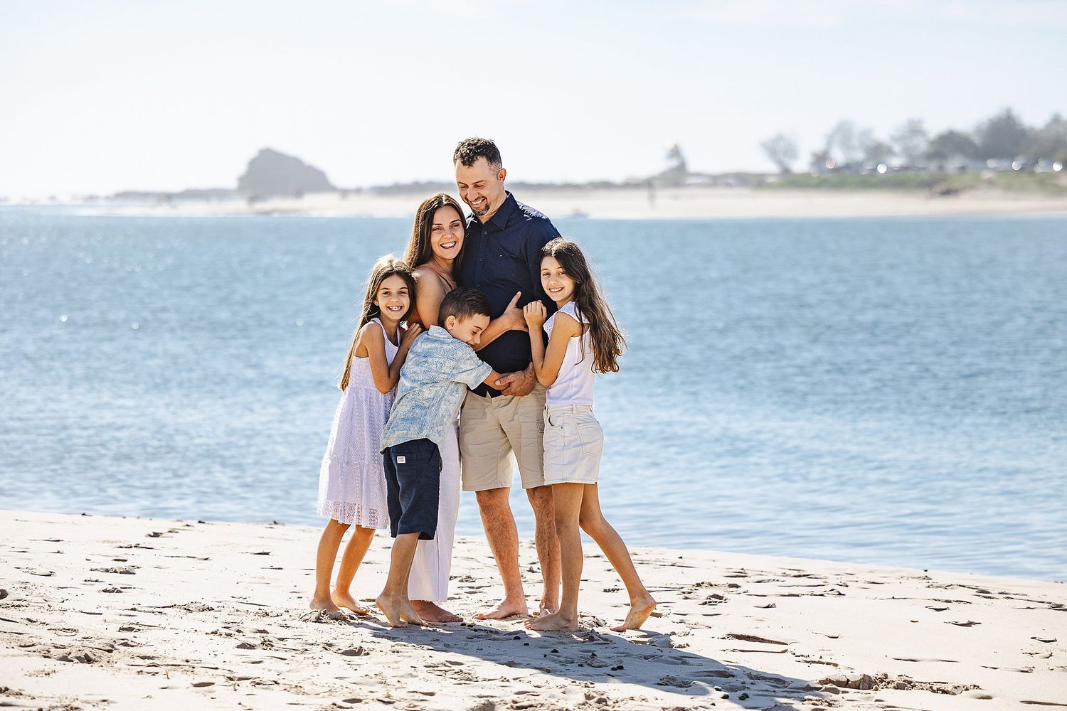 A happy family standing together and embracing on a sunny beach with blue ocean water and coastline in background.
