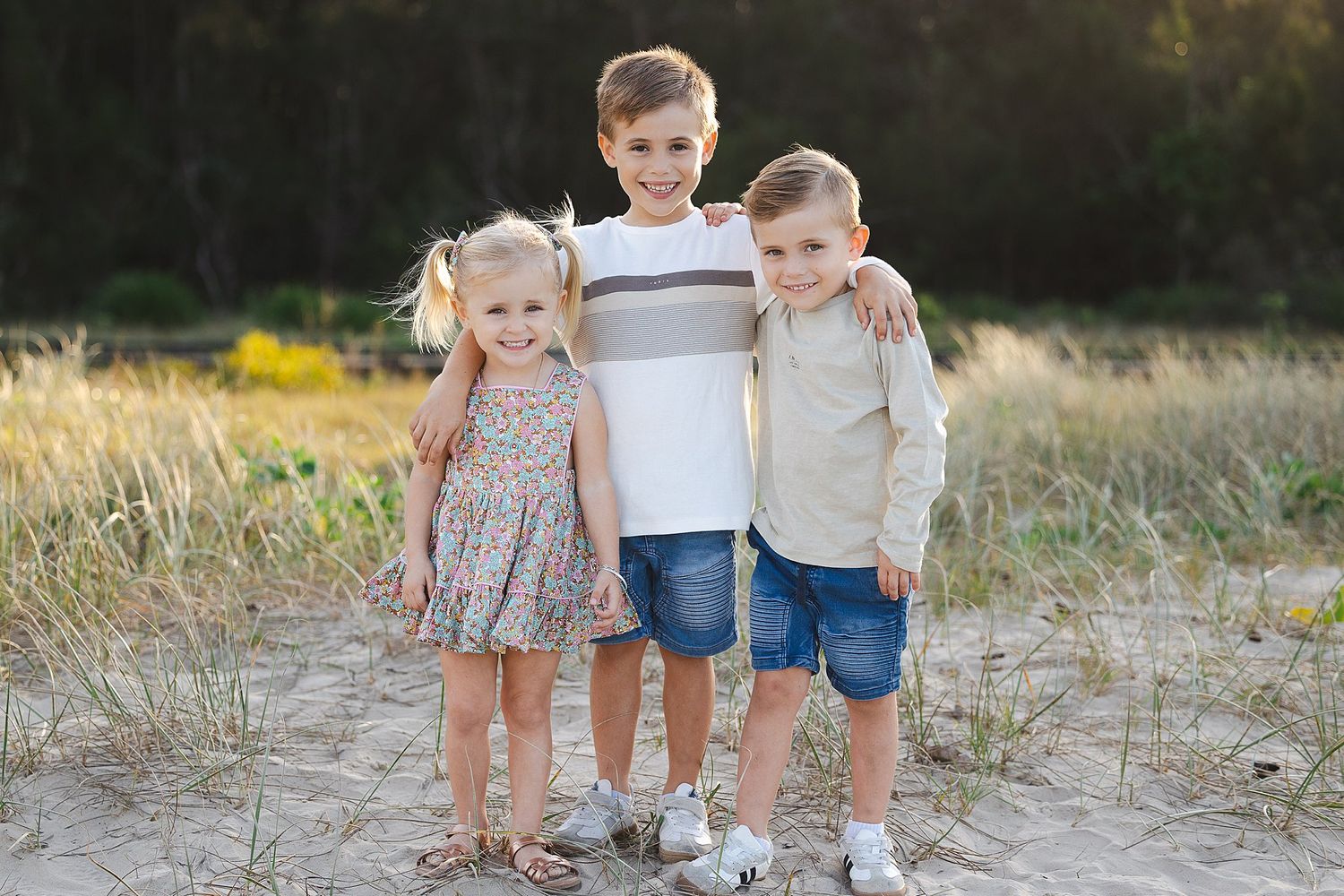Three young children posing together outdoors in a field with grass and sunlight in the background.