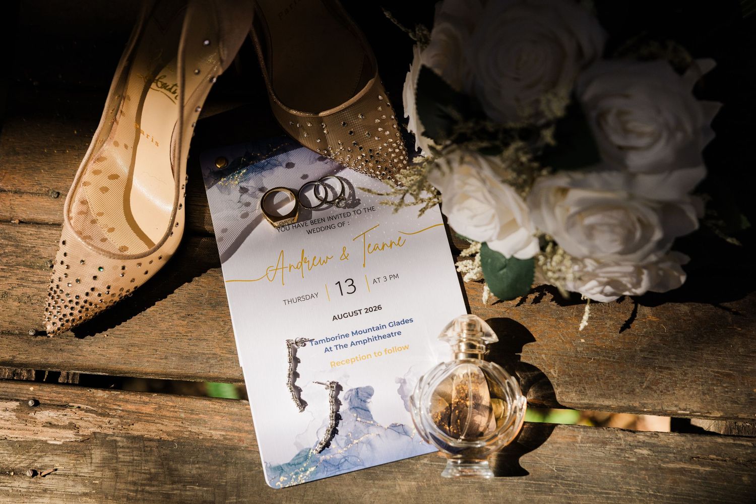Elegant wedding flat lay featuring Christian Louboutin heels, floral bouquet, invitation, and perfume on rustic wood.