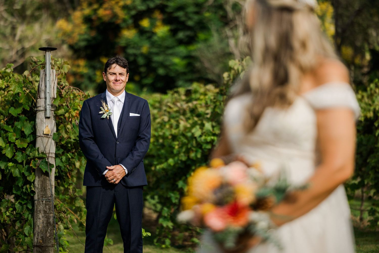 Groom in navy suit smiles at bride holding colorful bouquet during outdoor wedding ceremony surrounded by lush greenery.