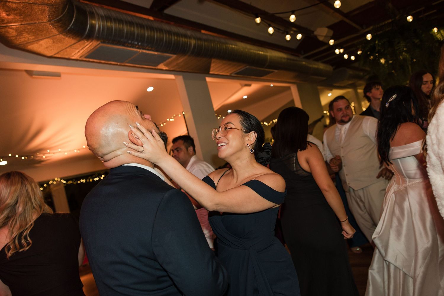 Couple dancing joyfully at a wedding reception, woman in navy dress smiling, warm ambient lighting in background.