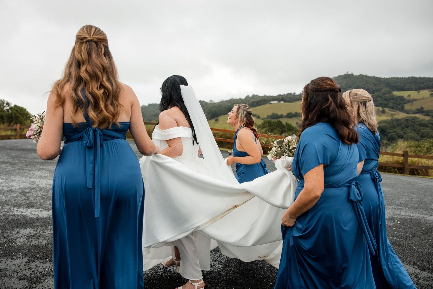 Bride and bridesmaids in blue dresses holding wedding veil outdoors with scenic hills backdrop.