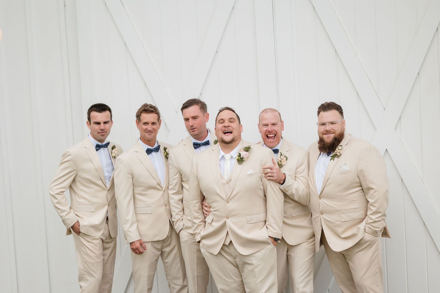 Six groomsmen in matching tan suits and blue bow ties pose together smiling at a wedding.