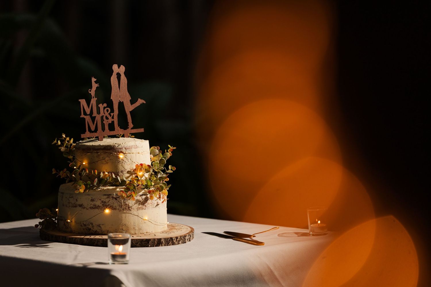 Two-tier wedding cake with Mr. & Mrs. topper on rustic wood slice, adorned with flowers and warm lights.