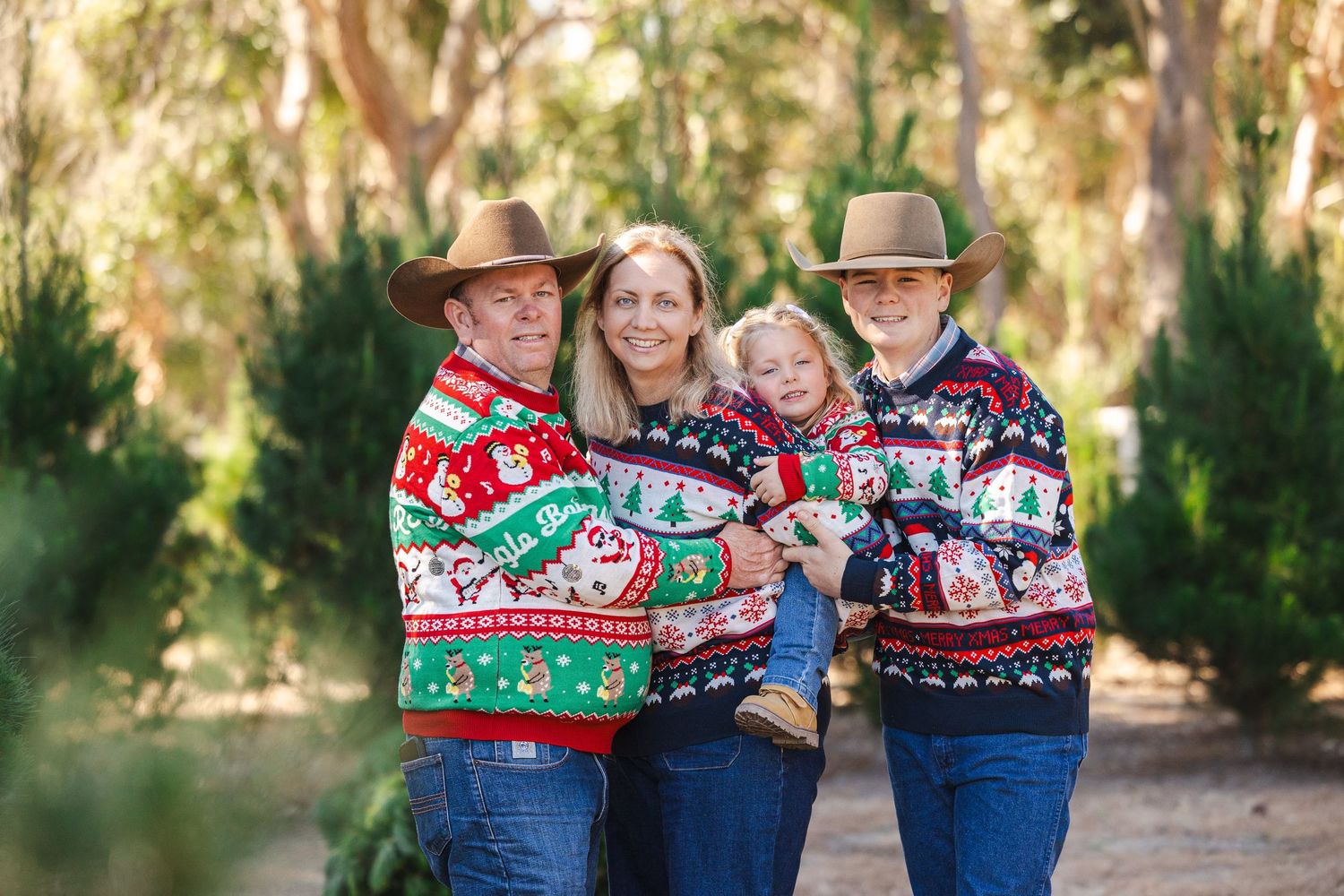 Three people wearing festive holiday sweaters pose together outdoors in a natural setting.