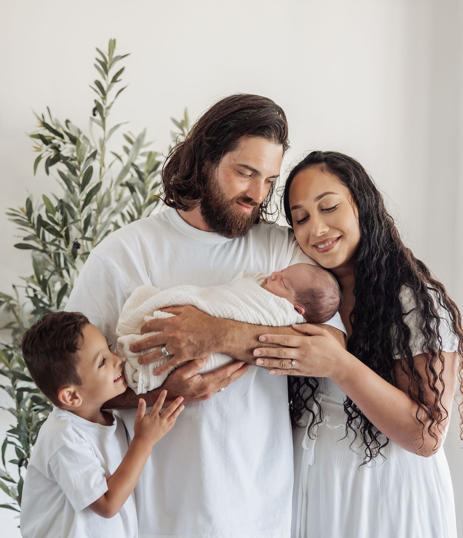 A family shares a tender moment together while holding their newborn baby near a green leafy plant.