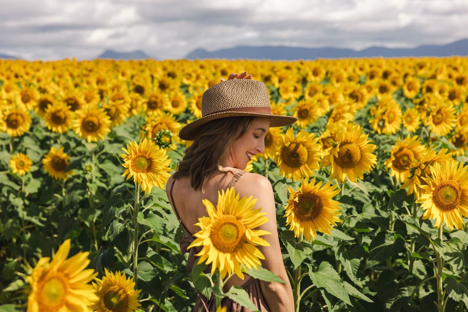 Woman in a hat standing among sunflowers in a large field with mountains in the background.