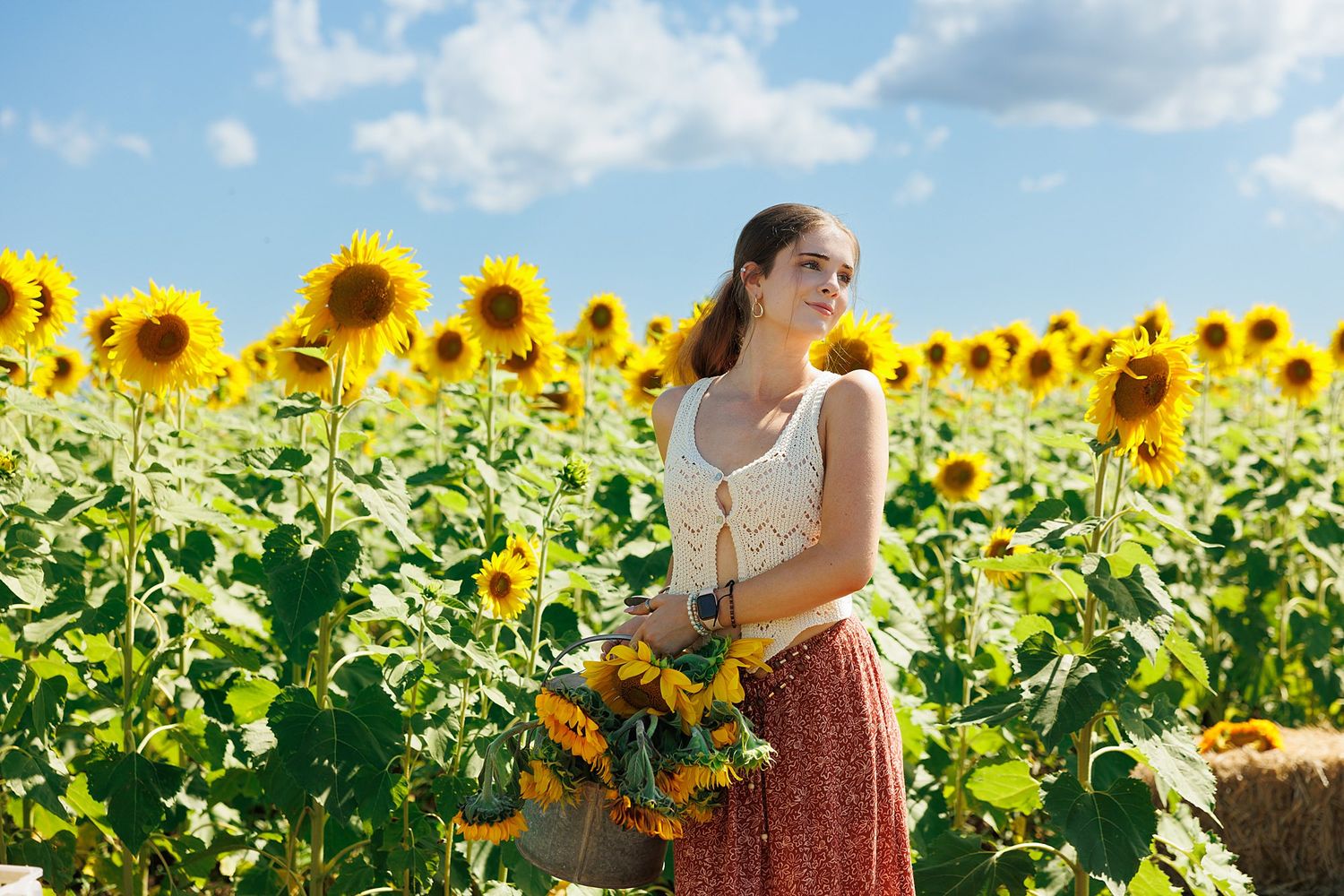A woman in a white tank top and floral skirt stands in a vibrant field of tall blooming sunflowers under blue summer skies.