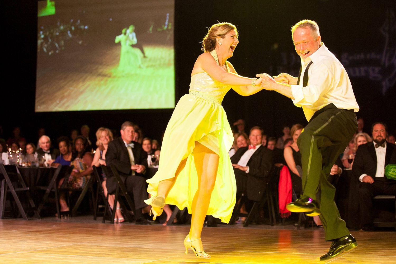 Dancers perform an energetic ballroom routine on stage as one partner spins in a flowing yellow dress.