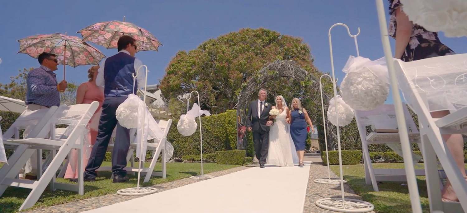 An outdoor wedding ceremony setup with white chairs and decorative umbrellas lining a pathway under blue skies.