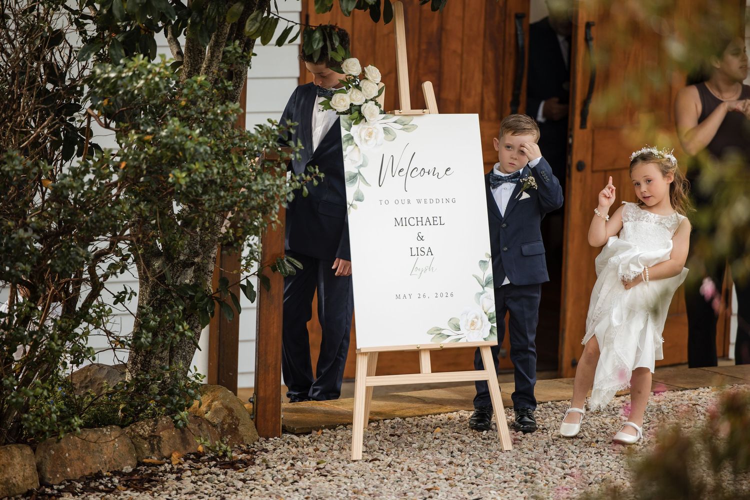Two children pose beside a wedding welcome sign for Michael and Lisa outdoors at a rustic venue.