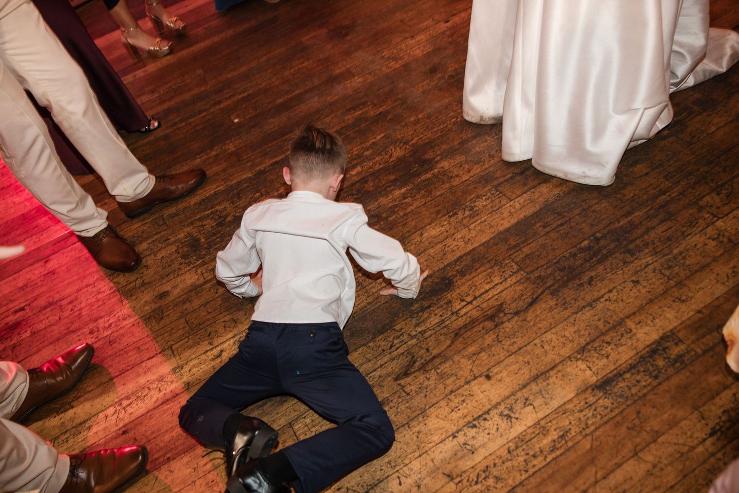 Young boy in dress clothes lying on wooden dance floor at a wedding reception.