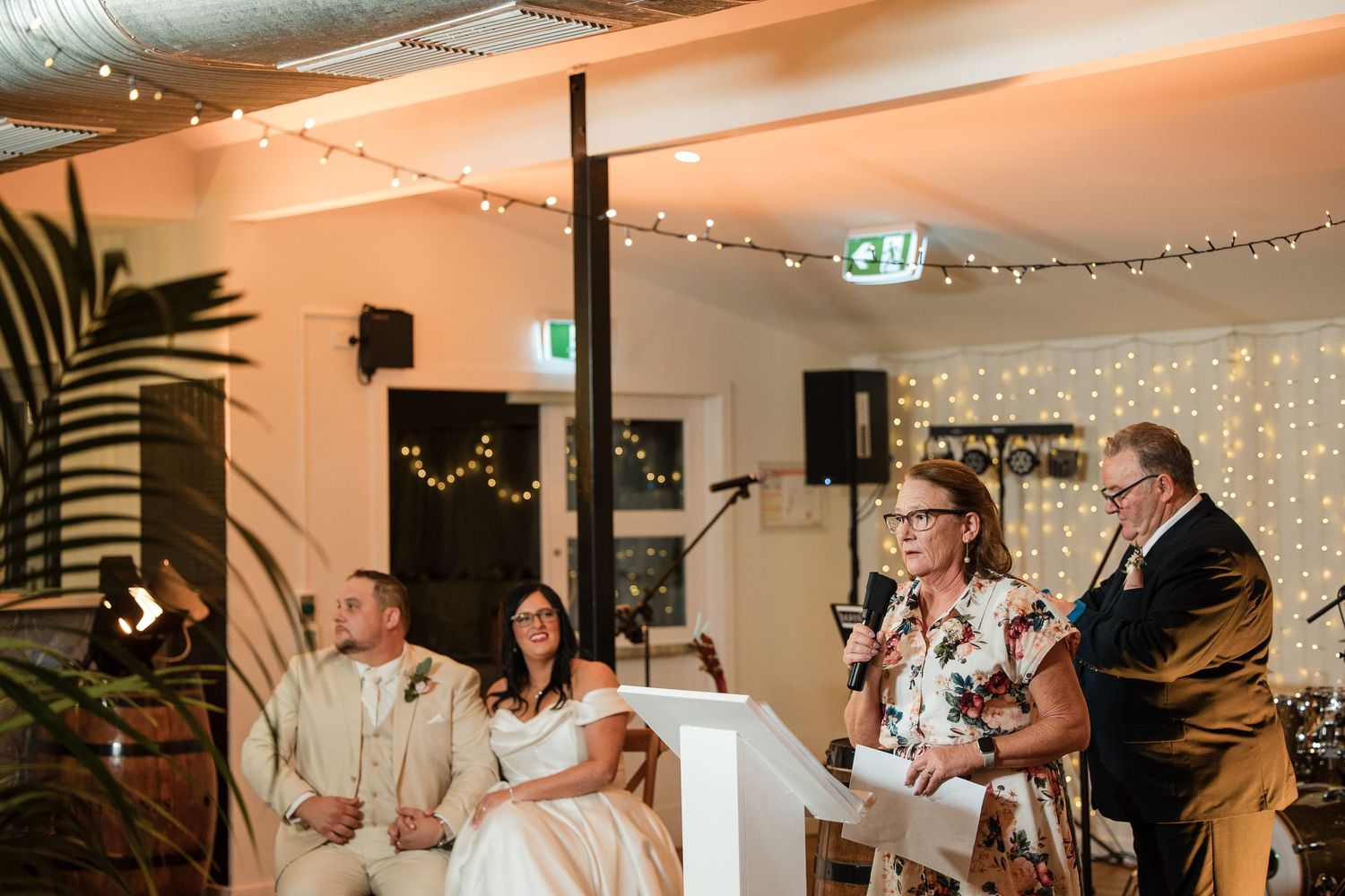 Couple listens as parents give a heartfelt wedding speech at an elegant reception venue decorated with fairy lights.