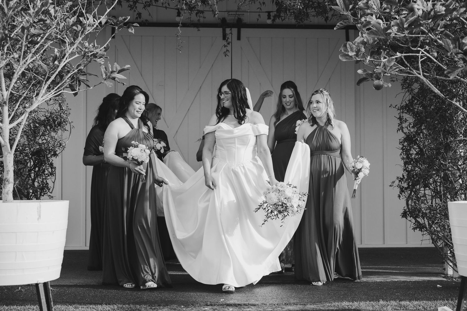 Bride in white gown walks with bridesmaids in gray dresses outside barn doors in black and white photo.