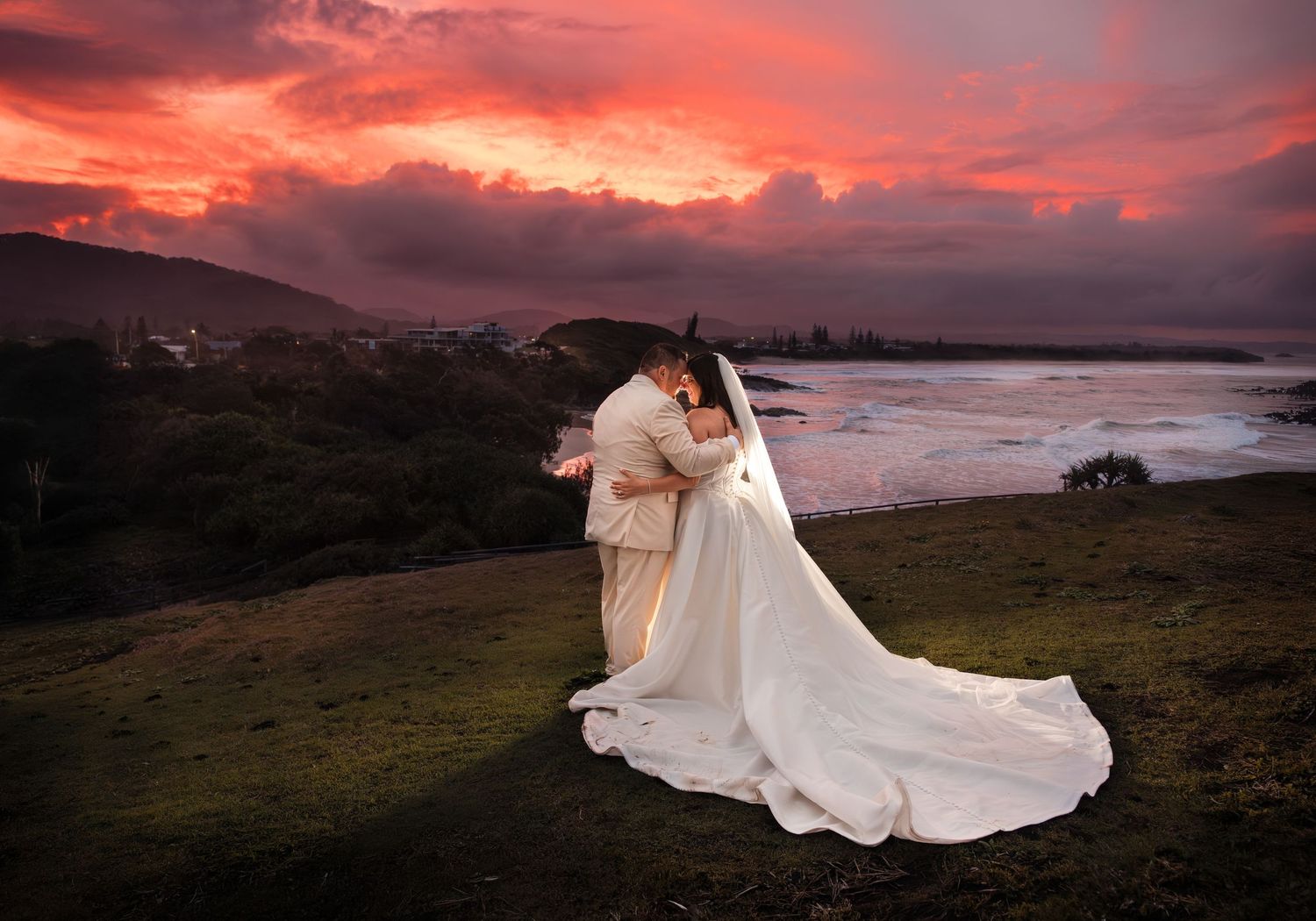 Bride and groom embracing at sunset with dramatic pink sky and coastal landscape backdrop.