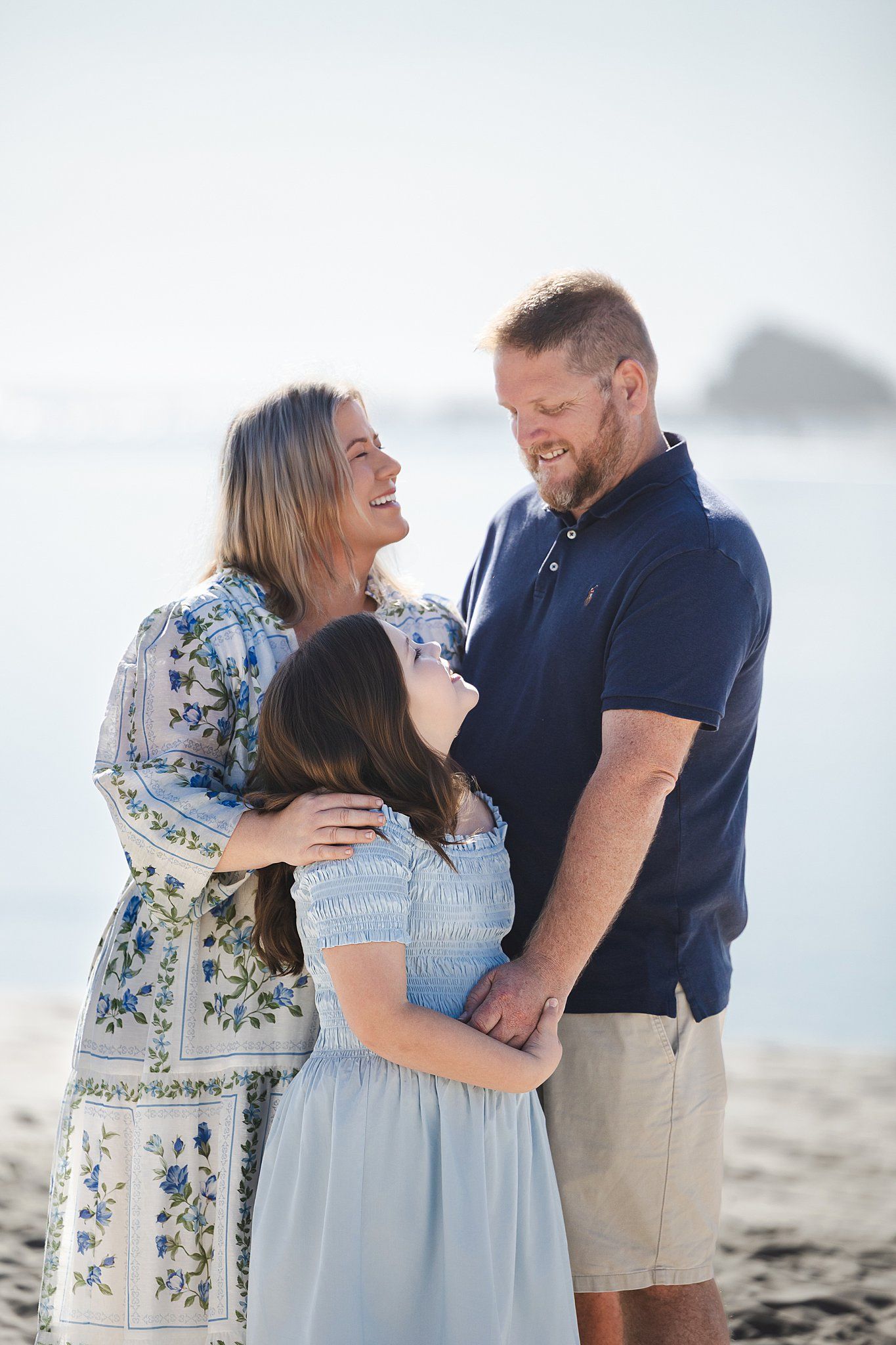 A happy family shares a joyful moment together while standing on a beach with ocean and mountains in the background.
