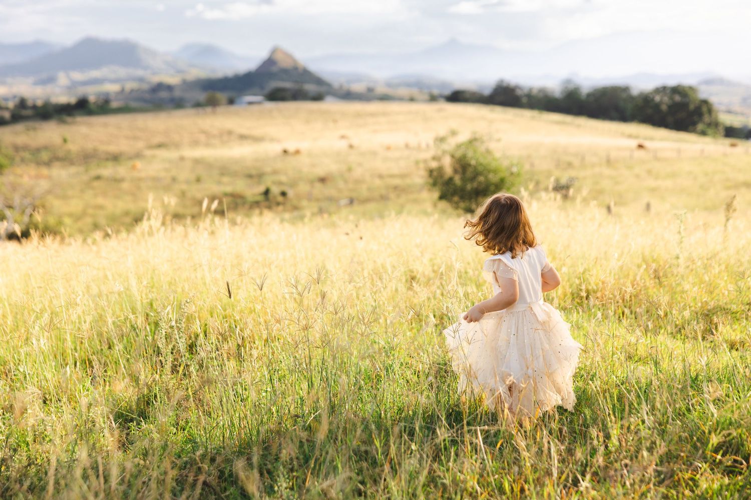 A person in a white dress runs through a golden sunlit meadow with mountains in the background at sunset.