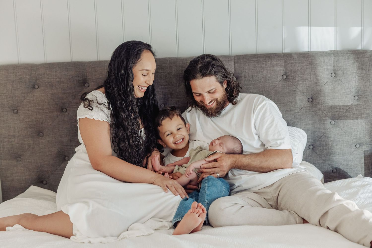 Happy family of four sitting on a bed, parents smiling at newborn baby with toddler child in cozy home setting.