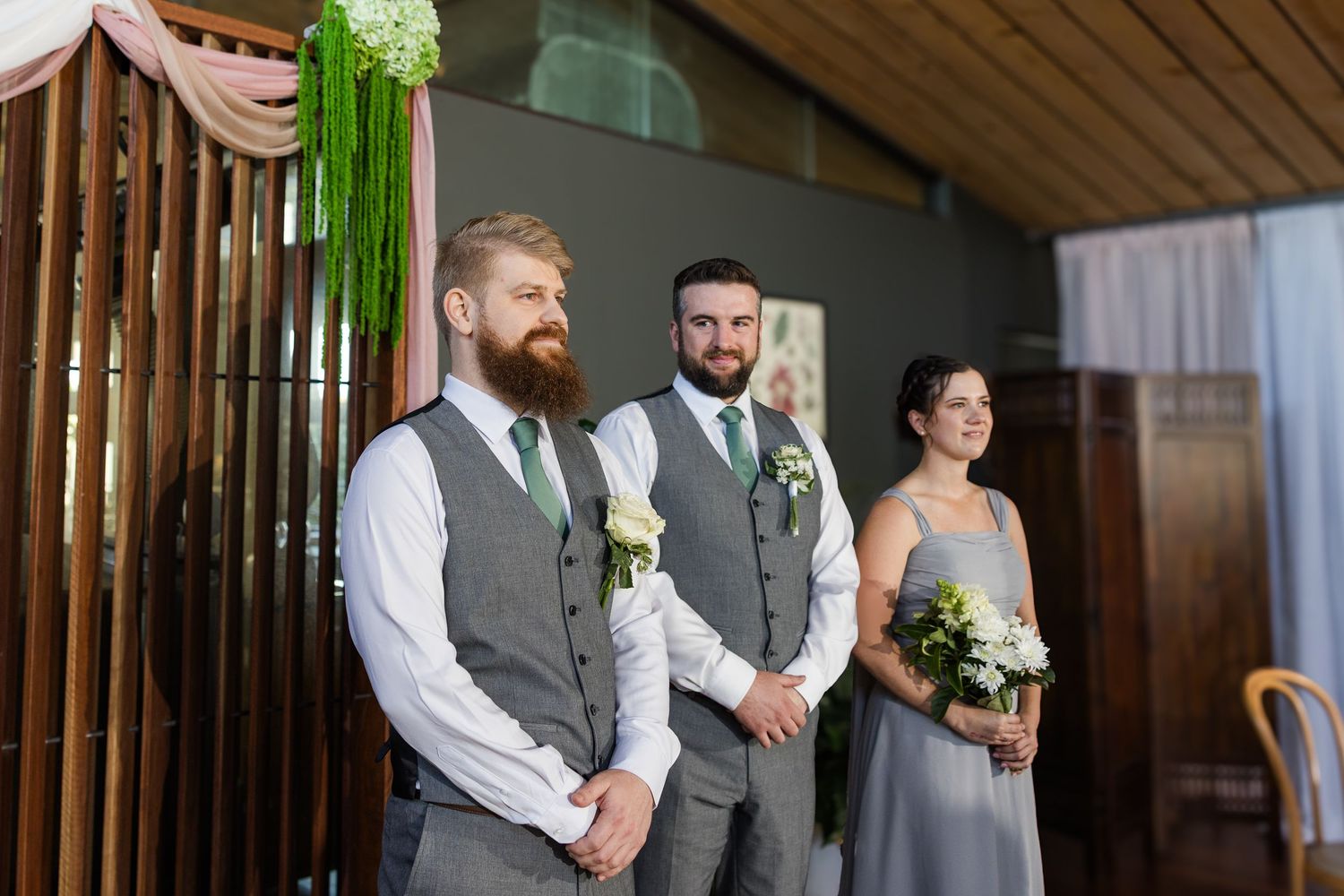Two groomsmen in gray vests and green ties stand with a bridesmaid holding flowers during a wedding ceremony.