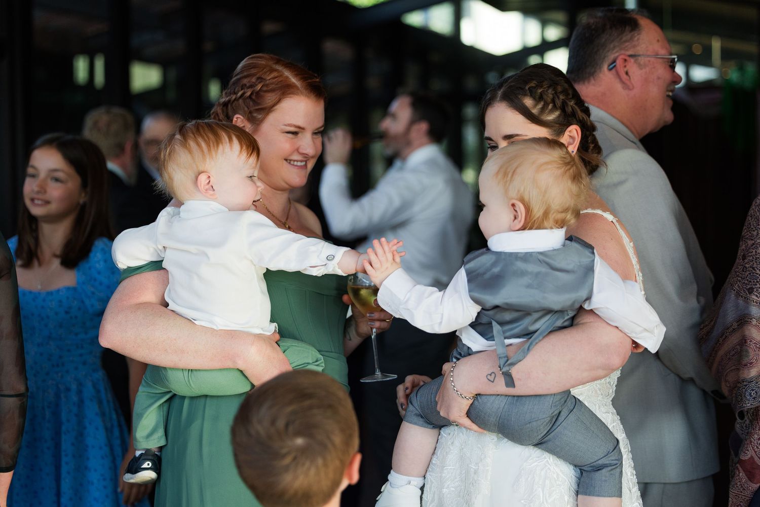 Two women in formal attire hold babies at a wedding reception, with guests socializing in the background.