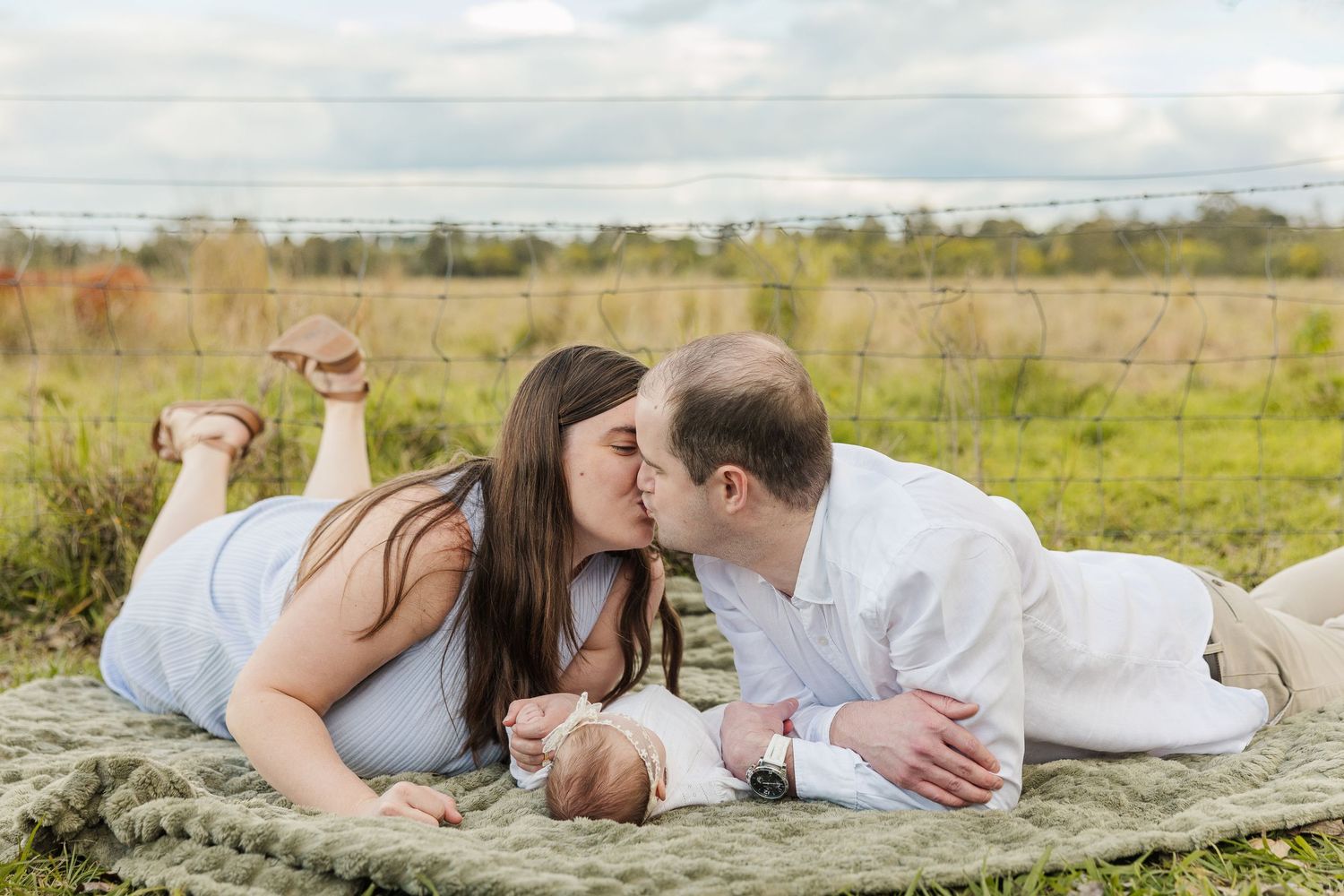 A couple kisses while lying on a blanket in a field with their newborn baby during an outdoor family photo session.