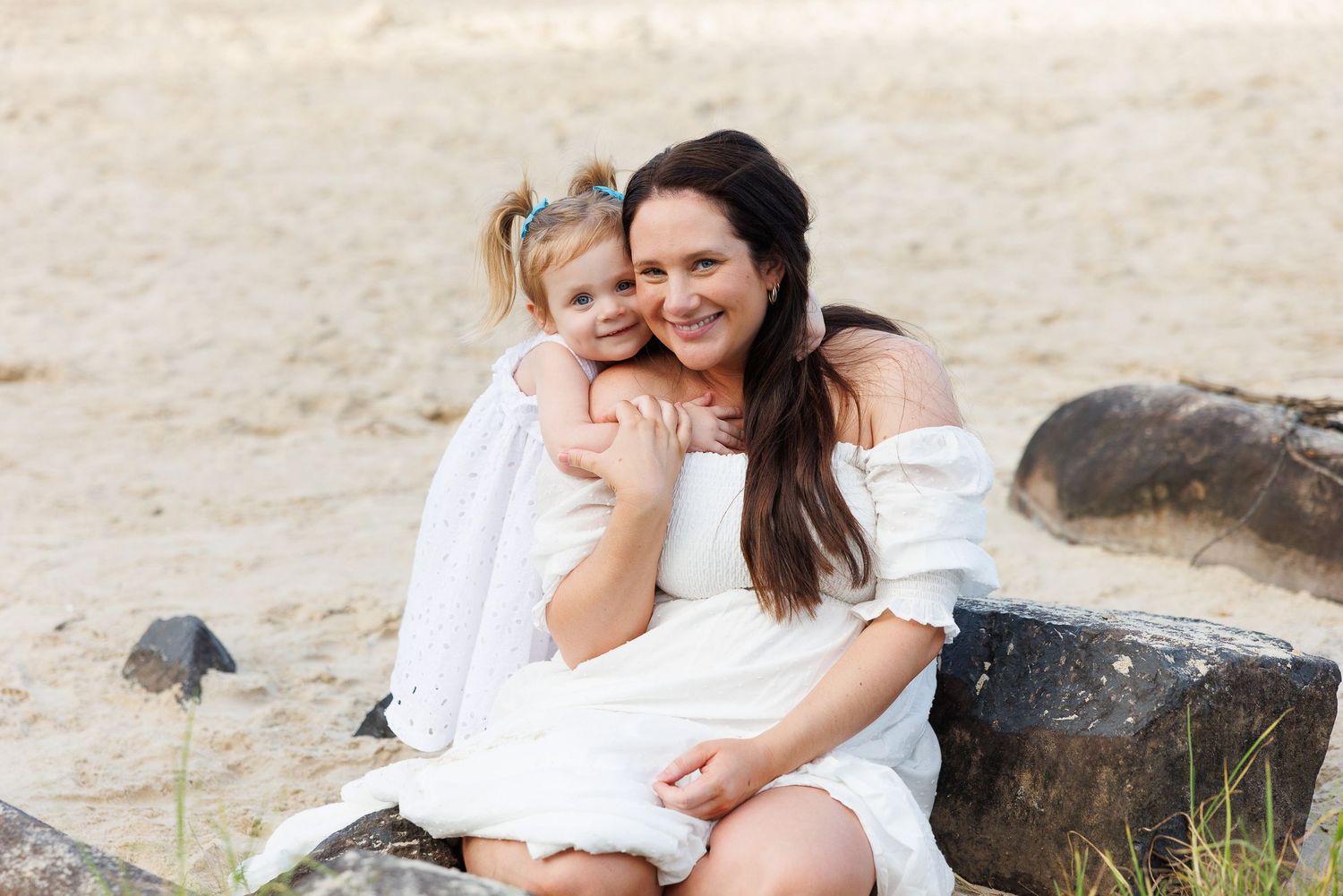 A woman and young girl in white dresses smile together on a sandy beach, sitting on rocks.