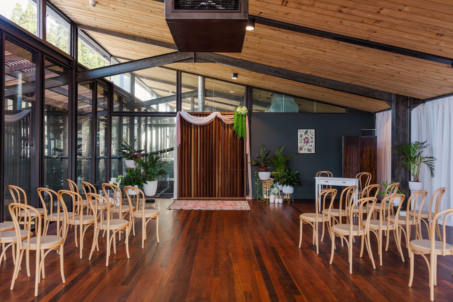 Elegant wedding ceremony venue with wooden chairs, hardwood floors, and a decorative backdrop surrounded by lush greenery.