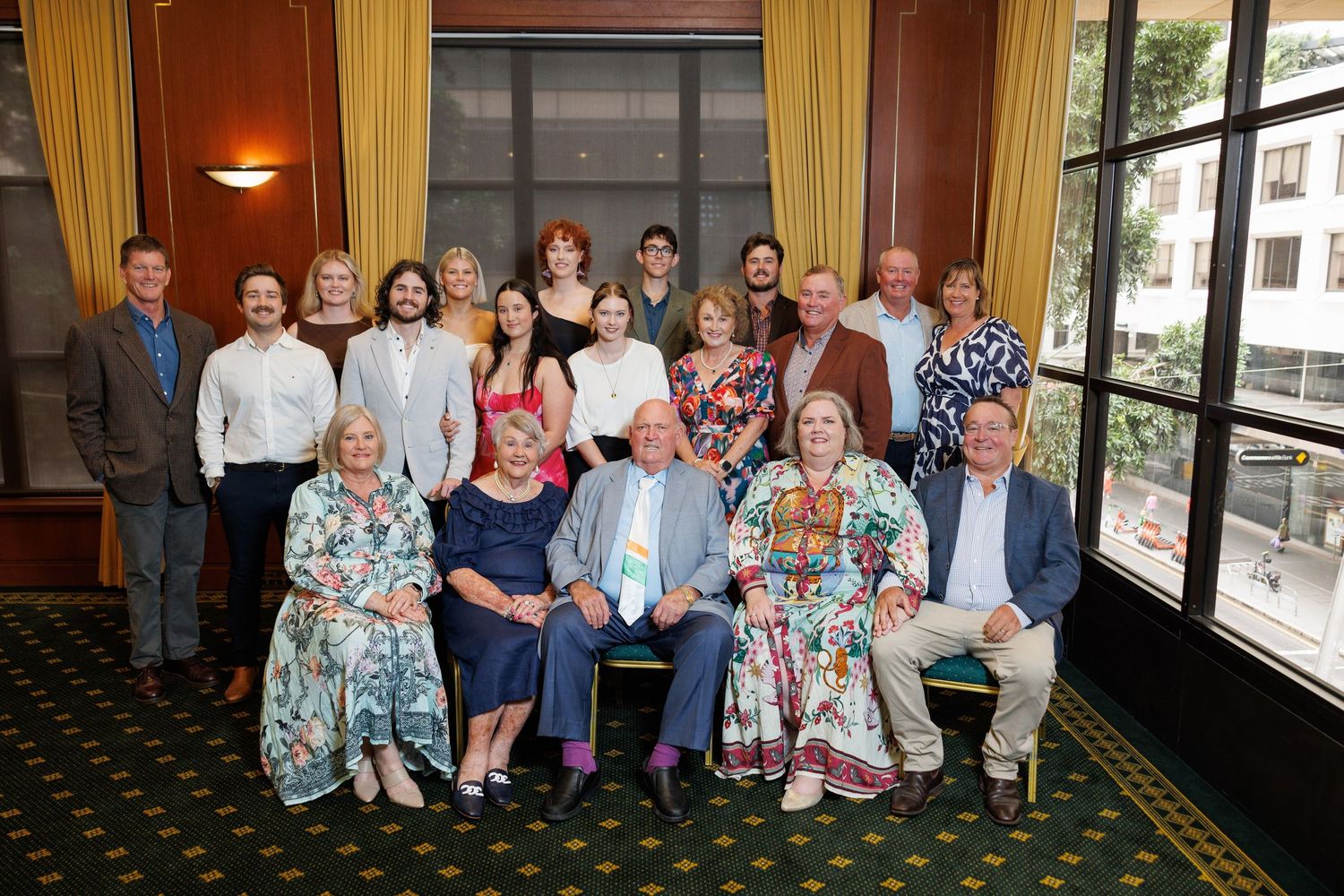 Group photo of people seated and standing together in a room with large windows and carpeted floor.