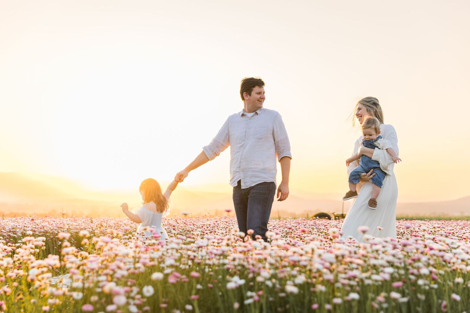 Family walking through a blooming flower field at sunset, parents holding child's hands while carrying a baby.
