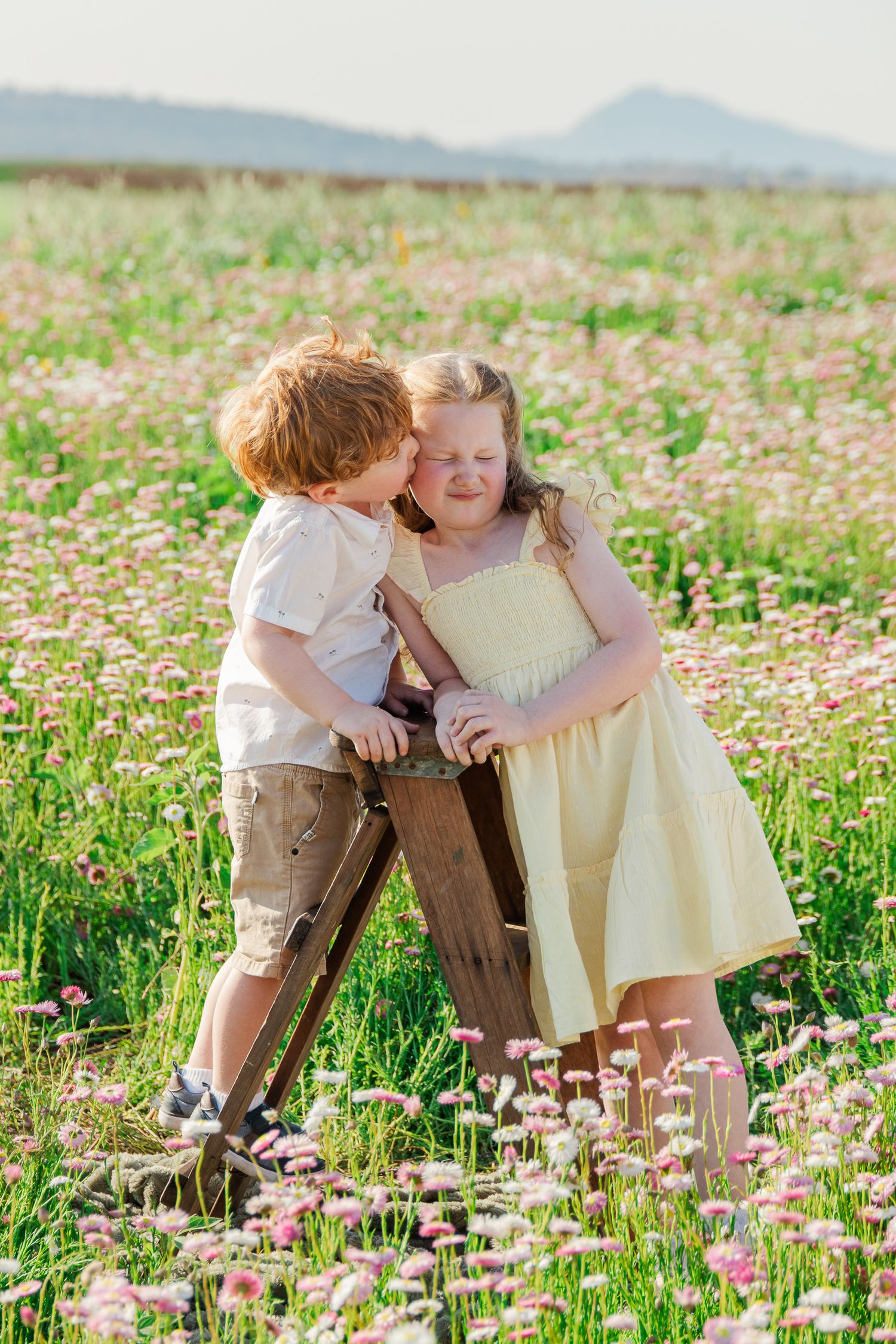 Two young children playing in a blooming flower field, a boy whispering to a girl in a yellow dress on a sunny day.
