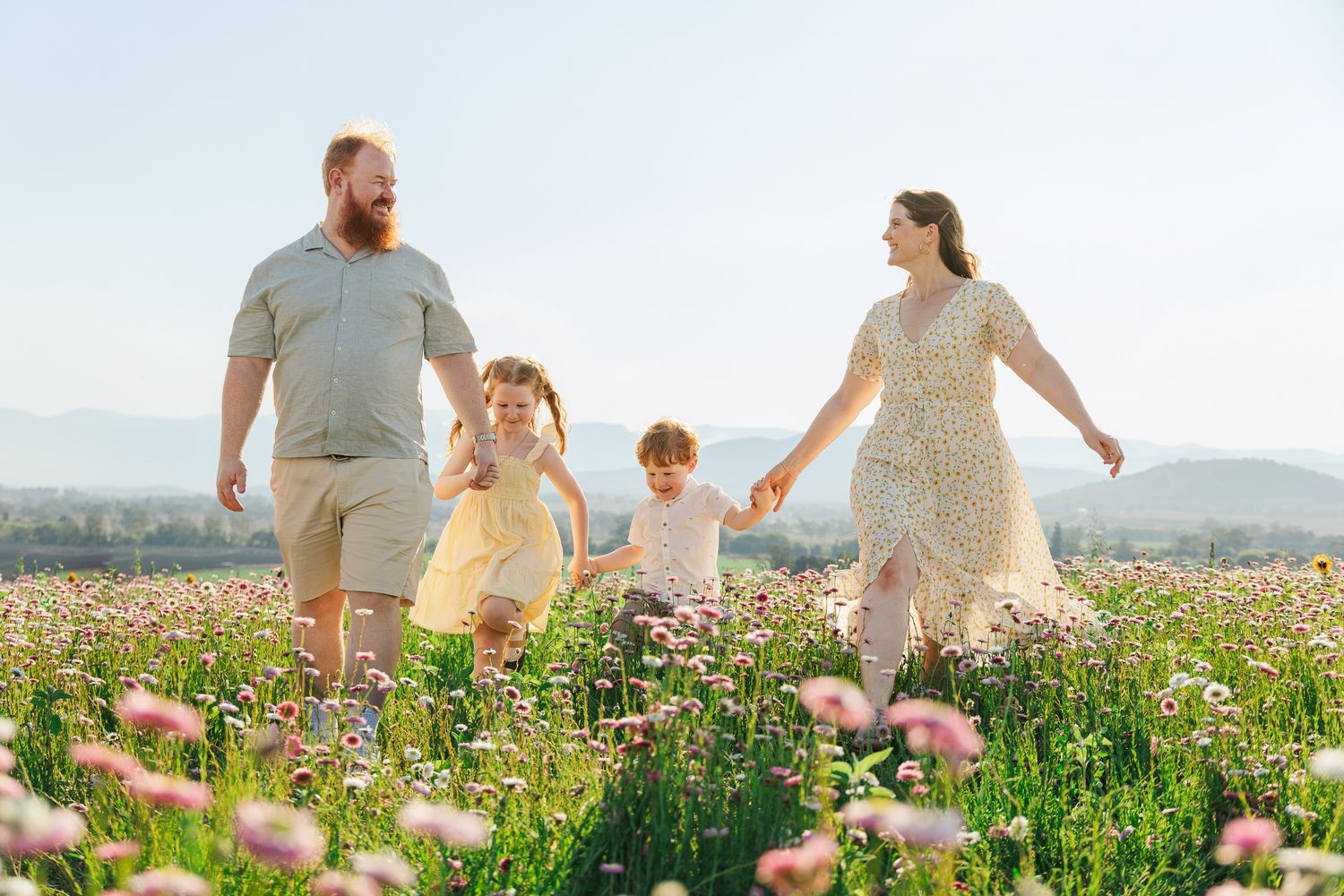 Family of four walking through a blooming wildflower field on a sunny day, with mountains in the background.