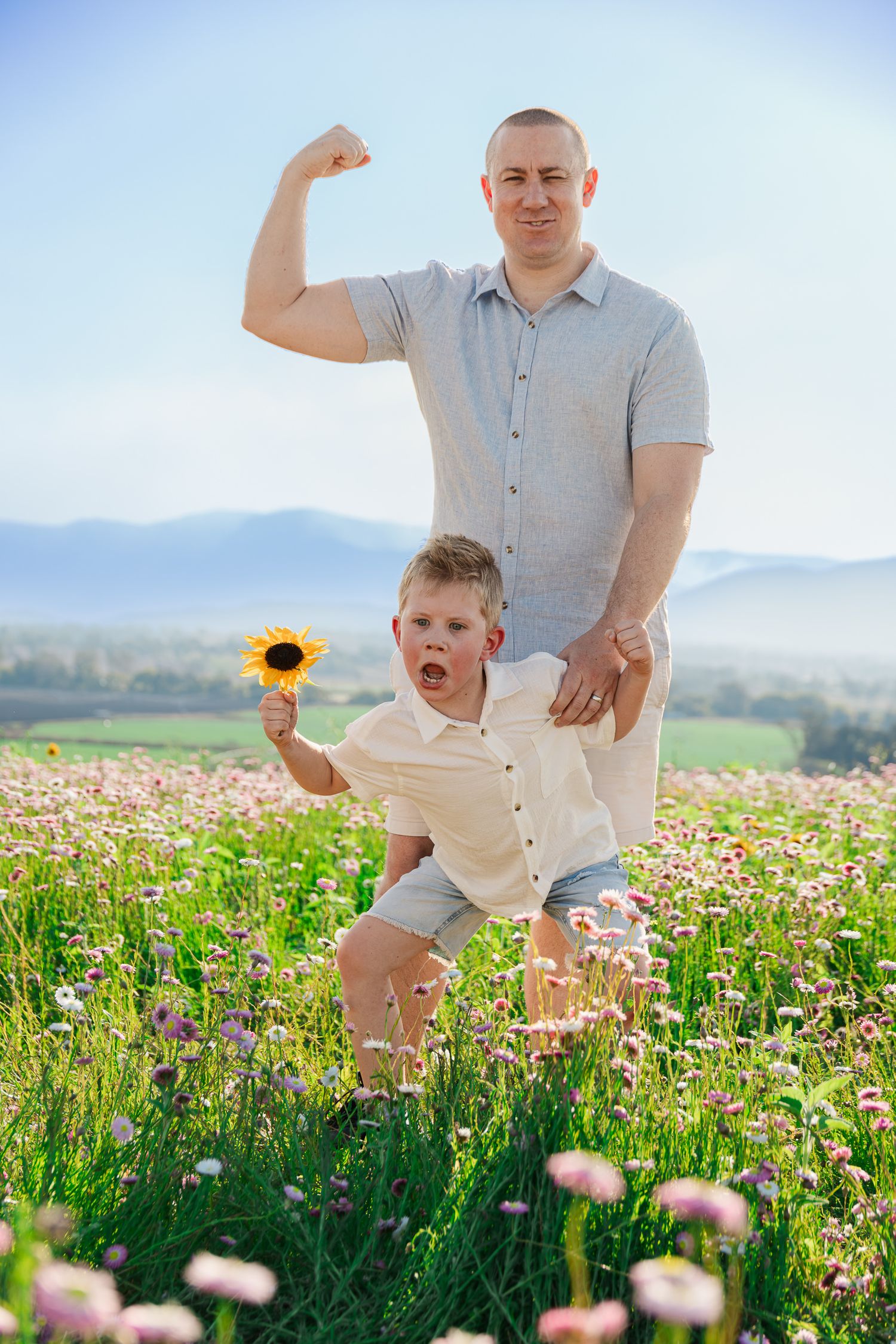 Father flexing muscles with son holding sunflower in blooming meadow with mountain backdrop.