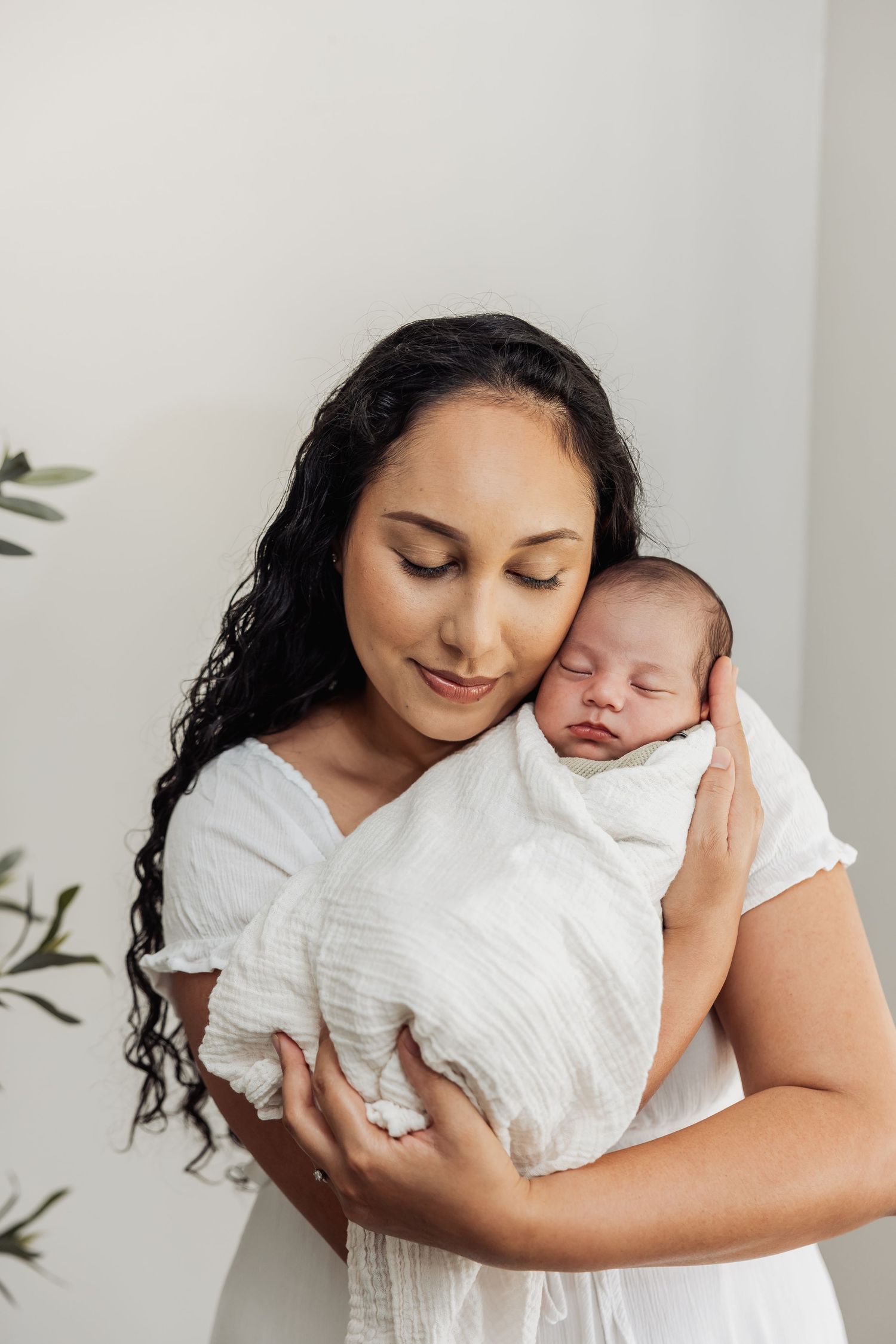 A mother in white dress tenderly holds her sleeping newborn baby wrapped in a white swaddle blanket.