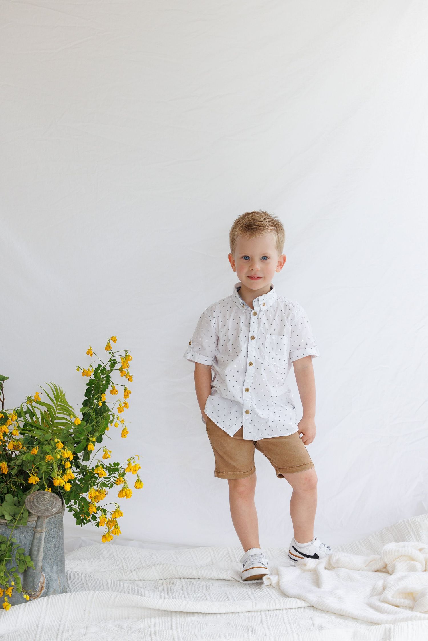 Young boy in white patterned shirt and tan shorts standing near yellow wildflowers against white backdrop.