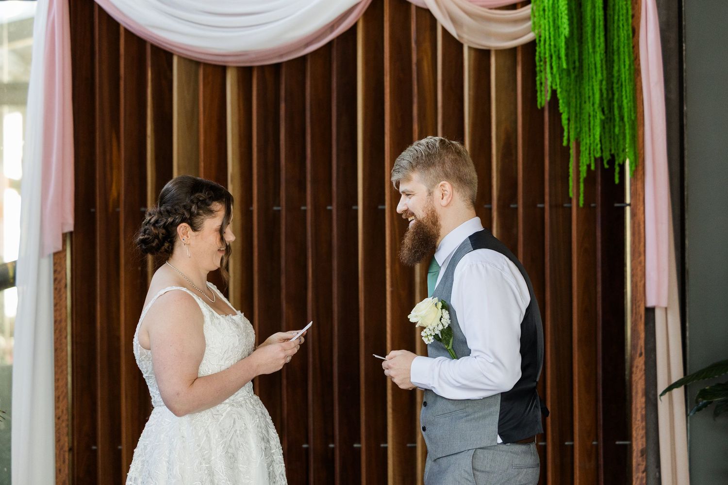 Bride and groom exchanging vows at wedding altar decorated with pink draping and green floral backdrop.