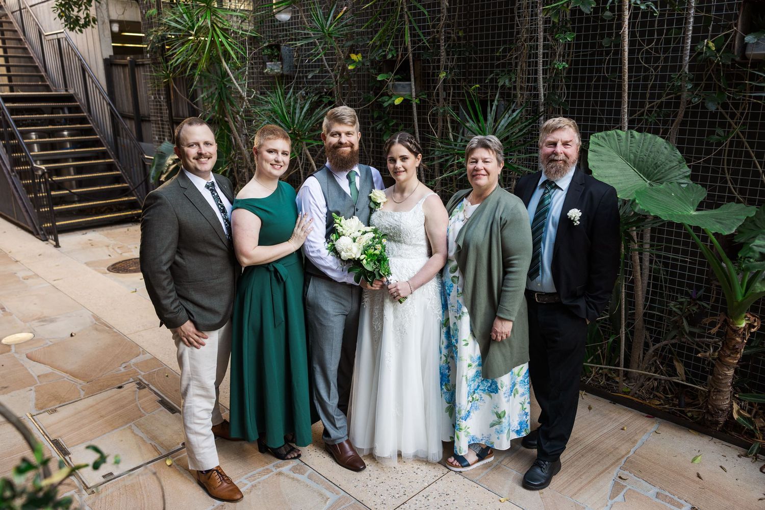 A wedding party of six poses indoors, featuring a bride in white, groom in gray vest, and guests in formal attire.