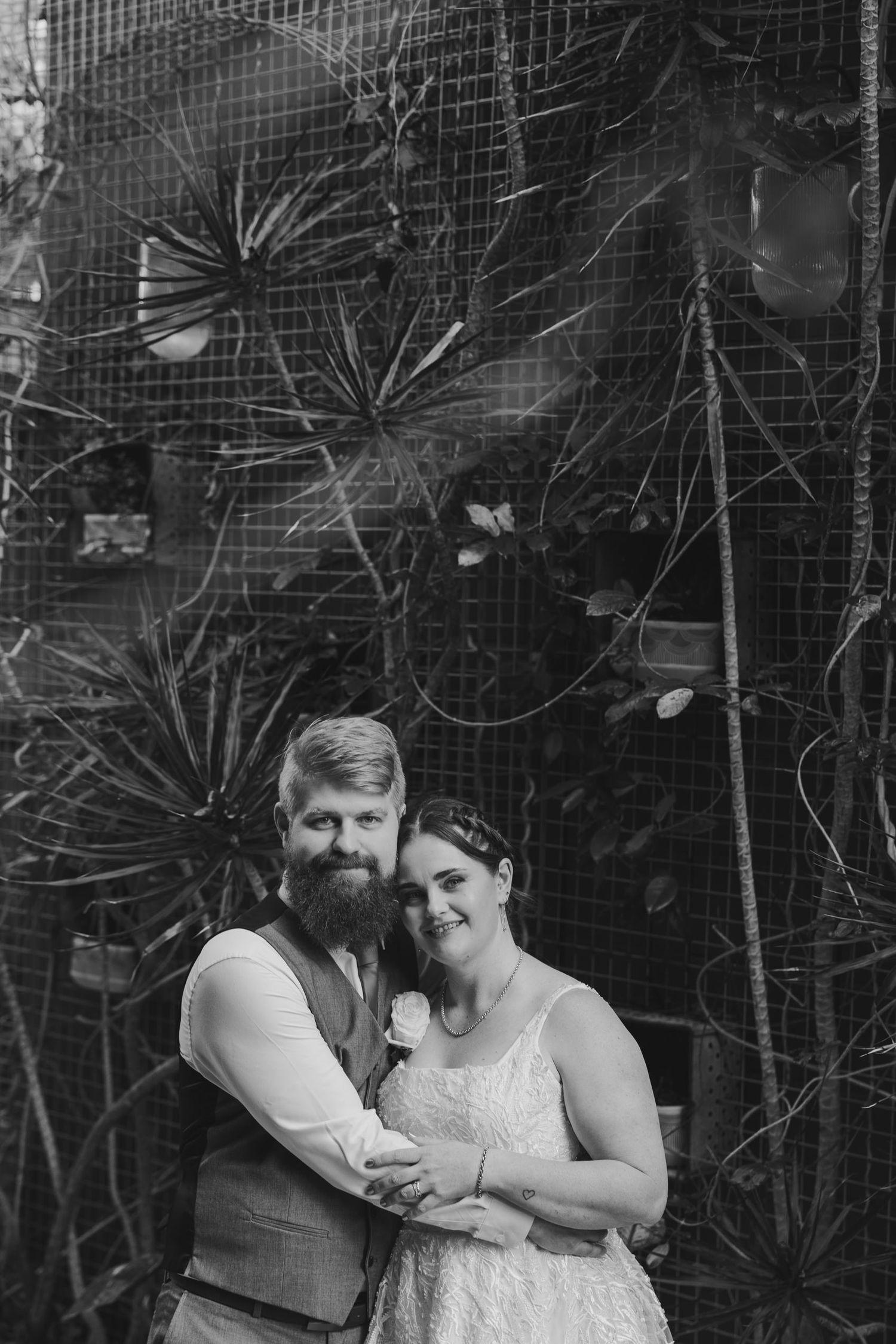 Black and white wedding portrait of a bearded groom and smiling bride embracing before a lush botanical backdrop.