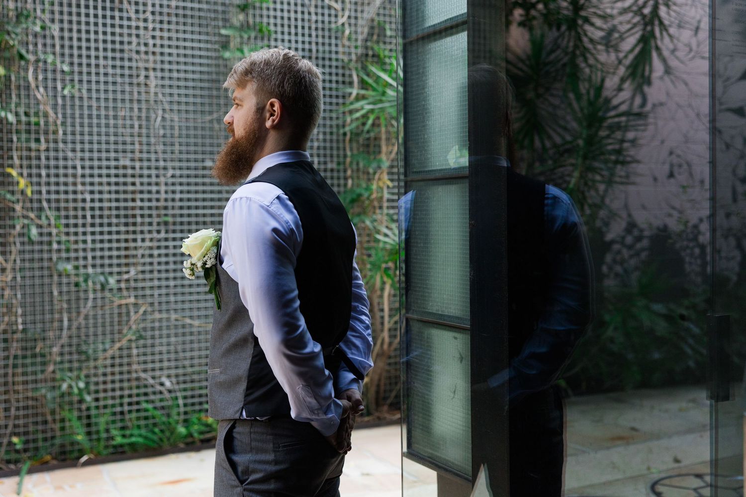 Bearded groom in gray vest holding white boutonniere, standing by glass door with green wall backdrop.