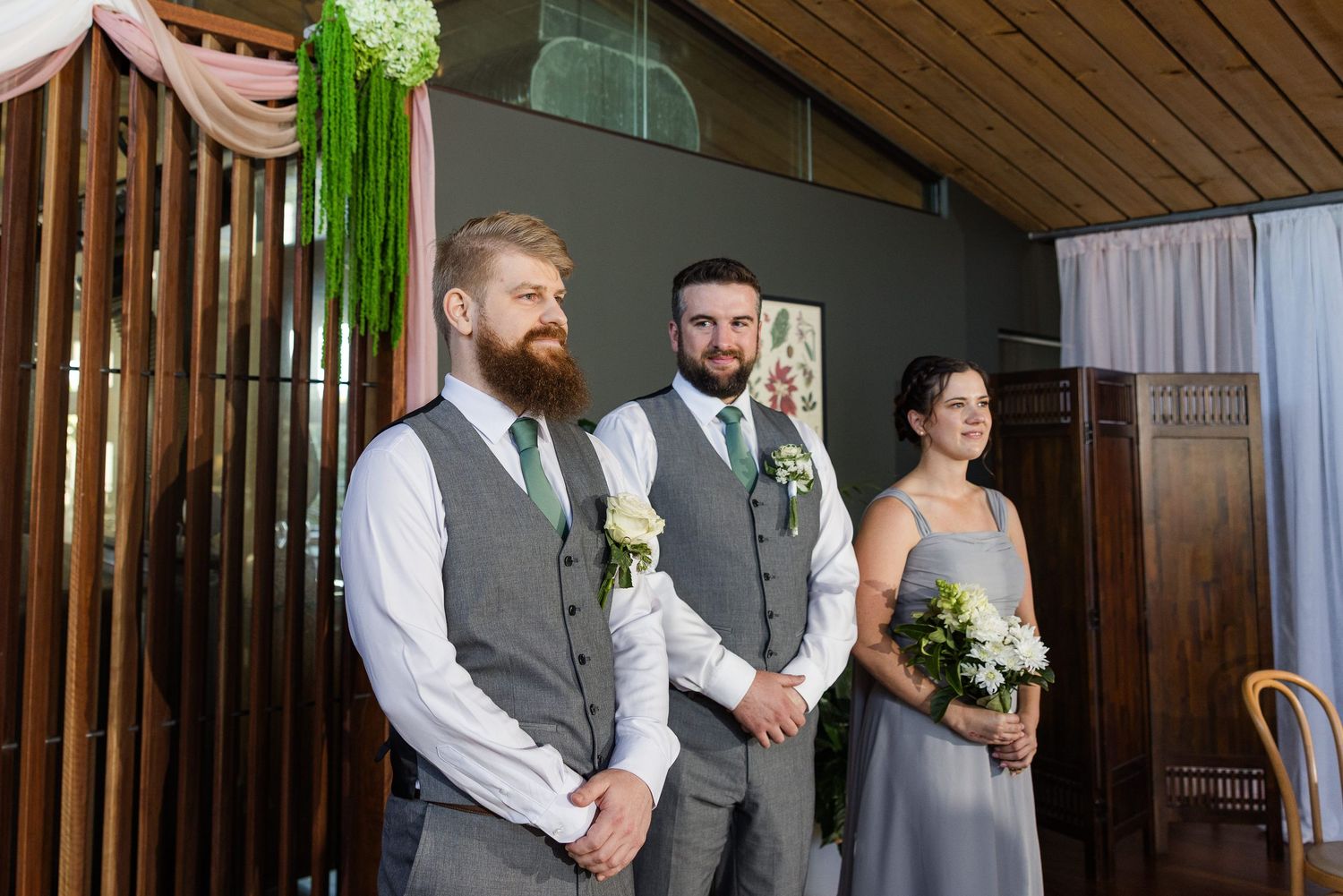 Two groomsmen in gray vests and green ties stand with a bridesmaid holding flowers at a wedding ceremony.