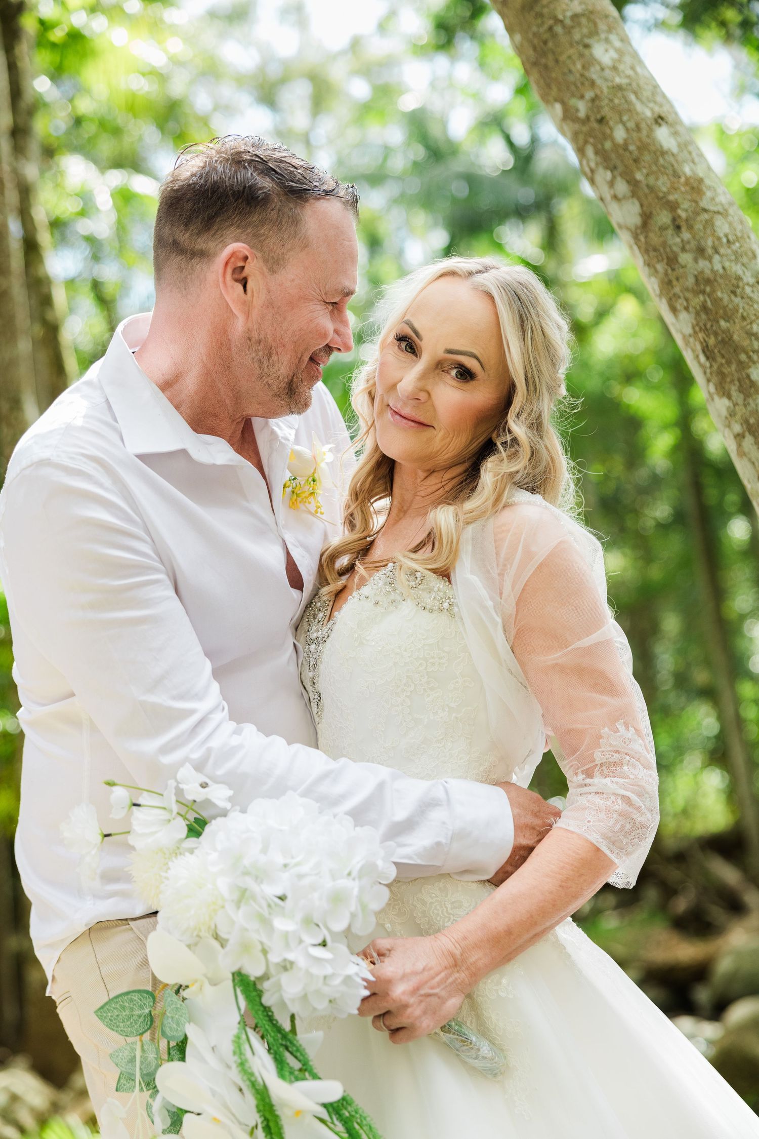 Couple in wedding attire embracing outdoors, bride holding white hydrangea bouquet, groom in white shirt with boutonniere.